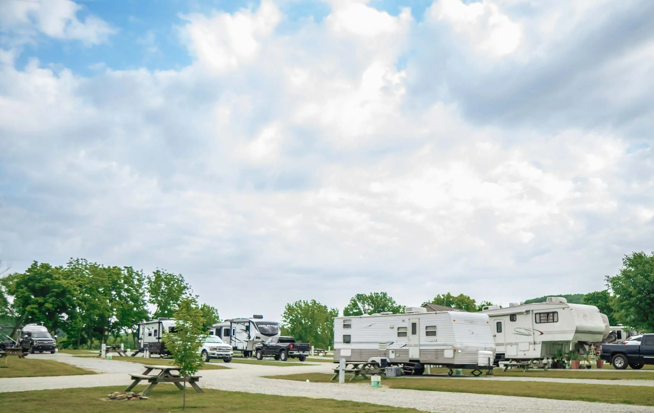 A campground with several RVs parked among trees, picnic tables, and a partly cloudy sky.