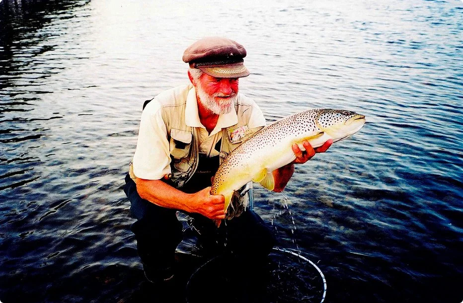 Man wearing a hat and beige shirt, holding a large rainbow trout fish over water.