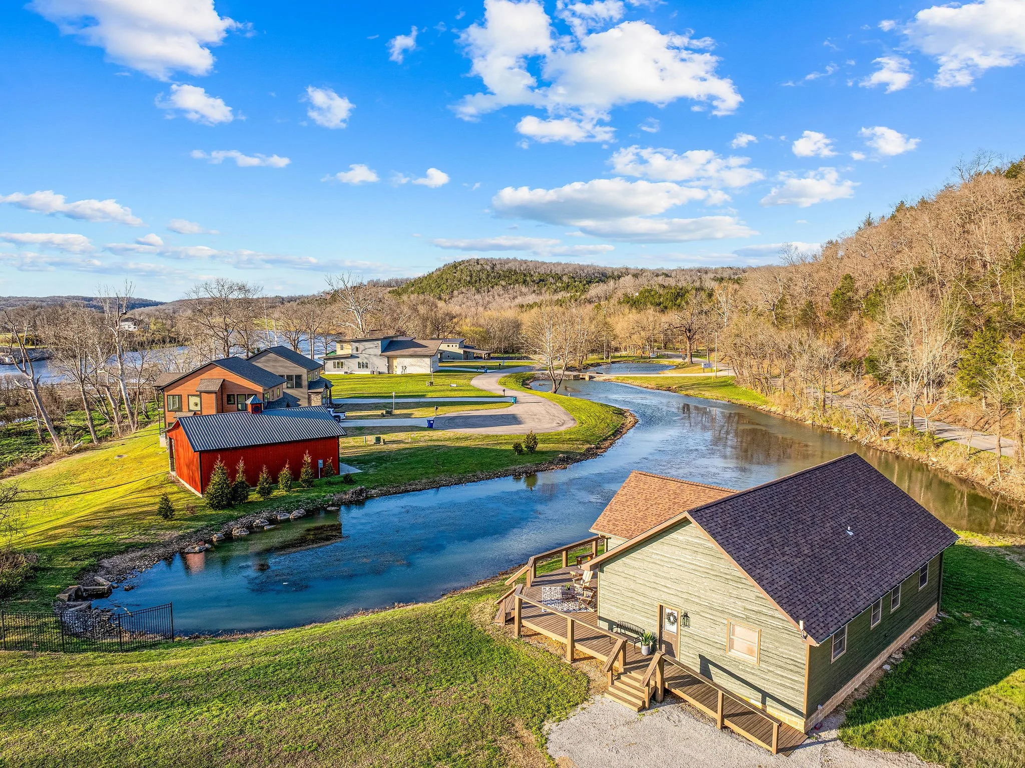A scenic view of a small town or neighborhood near a river, with houses, a road, trees, and hills under a bright blue sky with scattered clouds.