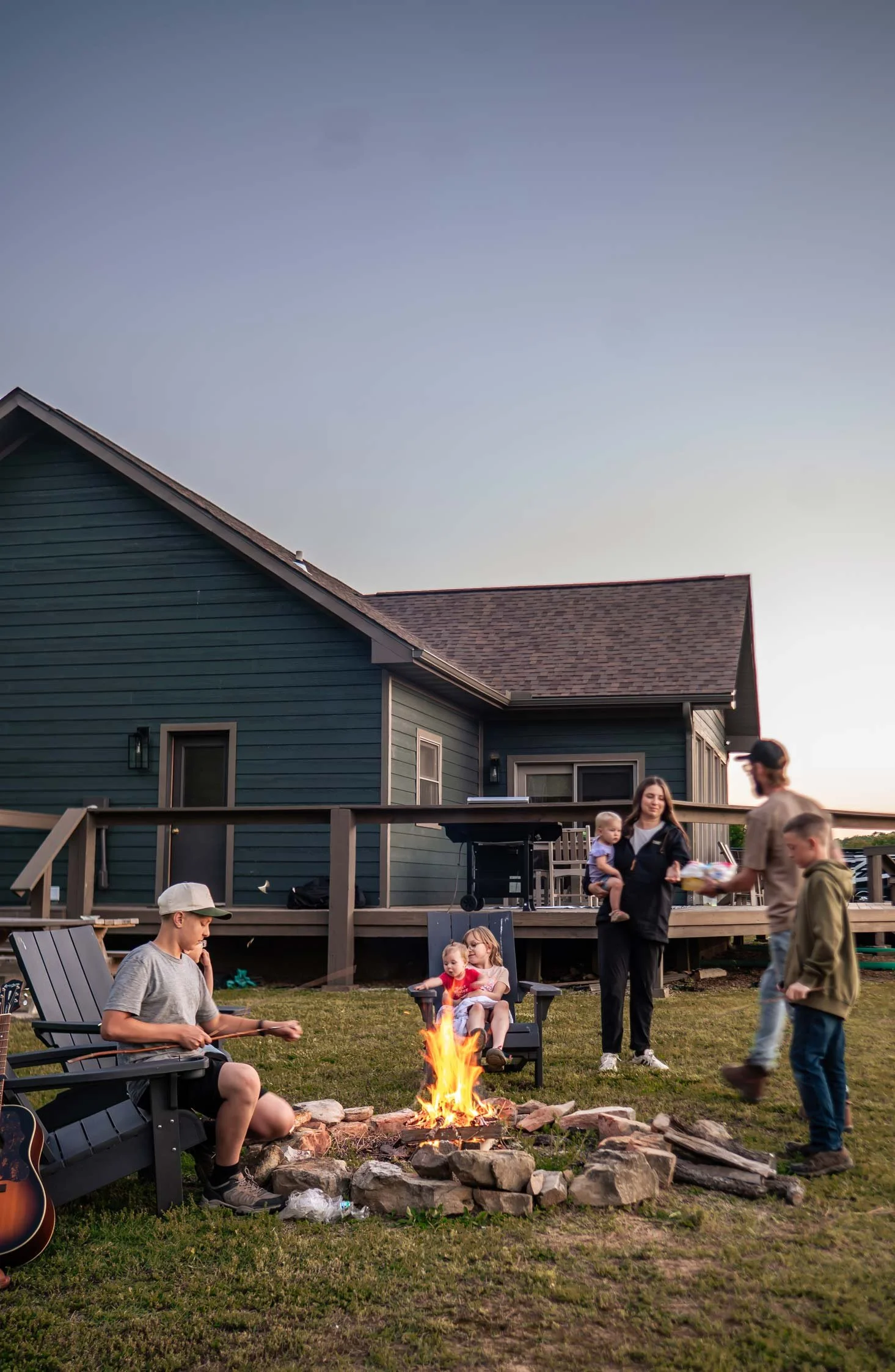 A group of people gathered around a campfire in a backyard at sunset, with a blue house and deck in the background.
