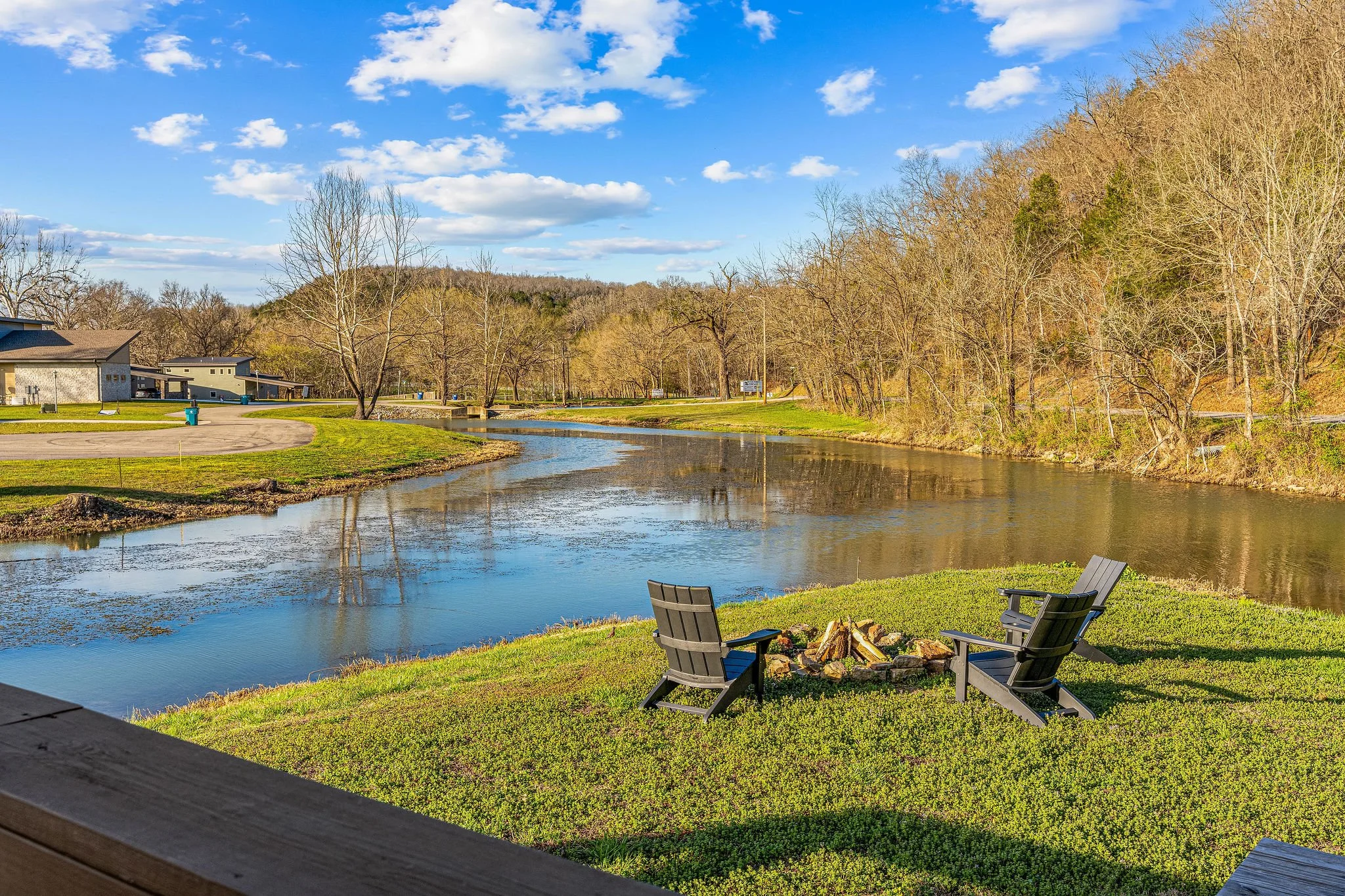 A peaceful riverside scene under a partly cloudy sky, with leafless trees, a grassy area, and wooden chairs arranged by the water, with some buildings in the background.