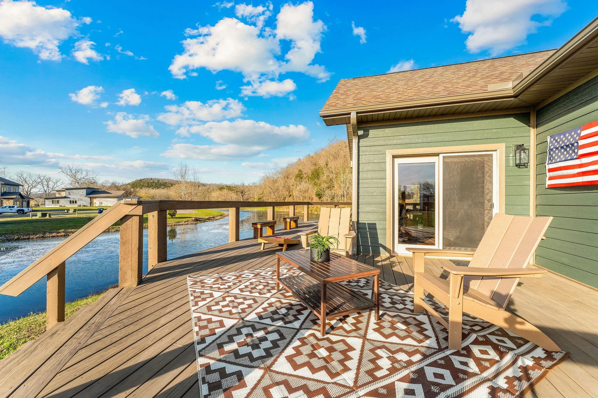 A wooden deck overlooking a river with modern houses in the background under a bright blue sky with scattered clouds. The deck has outdoor furniture, including chairs and a small table, with an American flag hanging on the house wall.