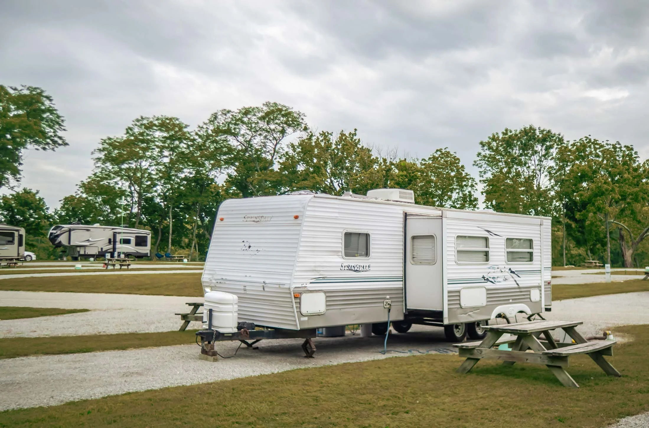 A white travel trailer parked on a gravel lot with a wooden picnic table in front, surrounded by grass and trees under a cloudy sky.