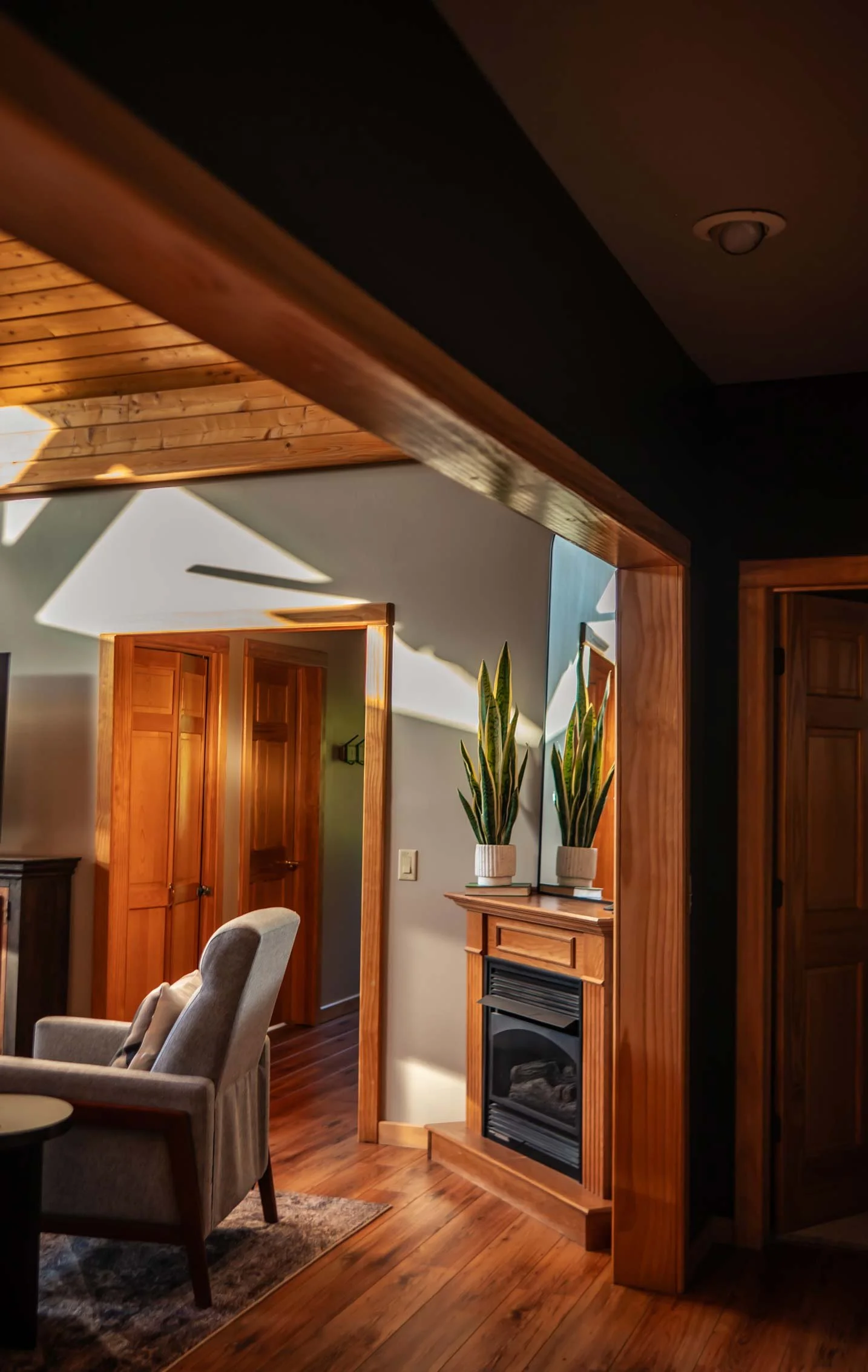 Interior of a room with wooden accents, a fireplace with a wooden mantel, two potted plants on a wooden stand, a gray armchair, and wood-paneled ceiling with sunlight casting shadows.