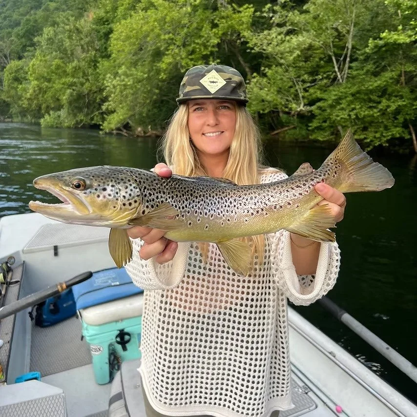 A woman wearing a camouflage hat and a white mesh sweater, smiling and holding a large fish, on a boat near a green, wooded shoreline.