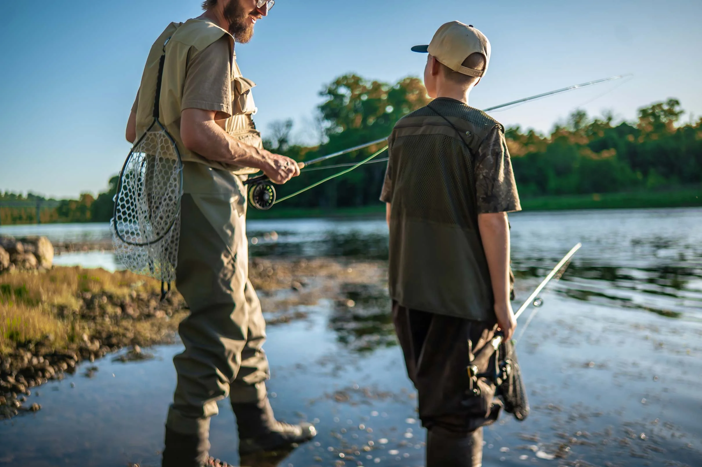 A man and a boy, both wearing fishing gear, standing by a body of water during the daytime. The man is showing the boy how to fish, holding a fishing rod and smiling at him. The scene has a clear, blue sky with trees in the background.