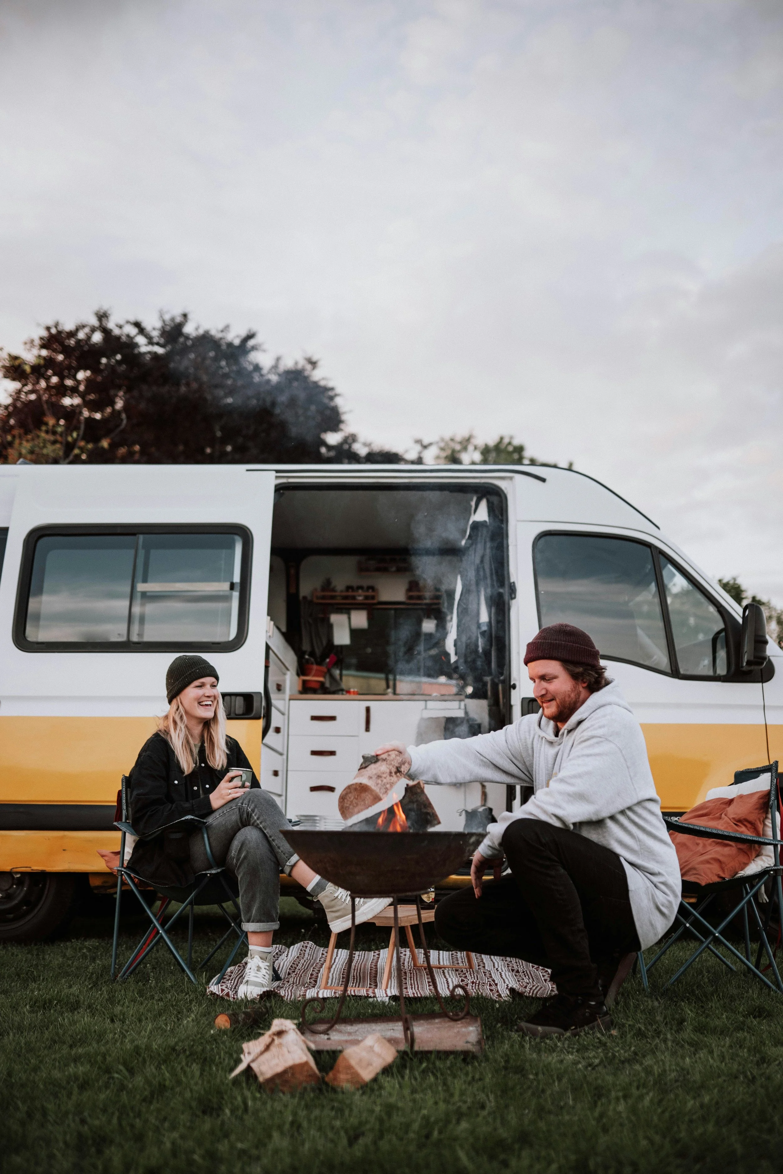 Two people camping outdoors beside a yellow and white camper van, sitting around a small fire with logs, and smiling. The woman is wearing a black beanie and jacket, and the man is wearing a gray hoodie and black beanie.