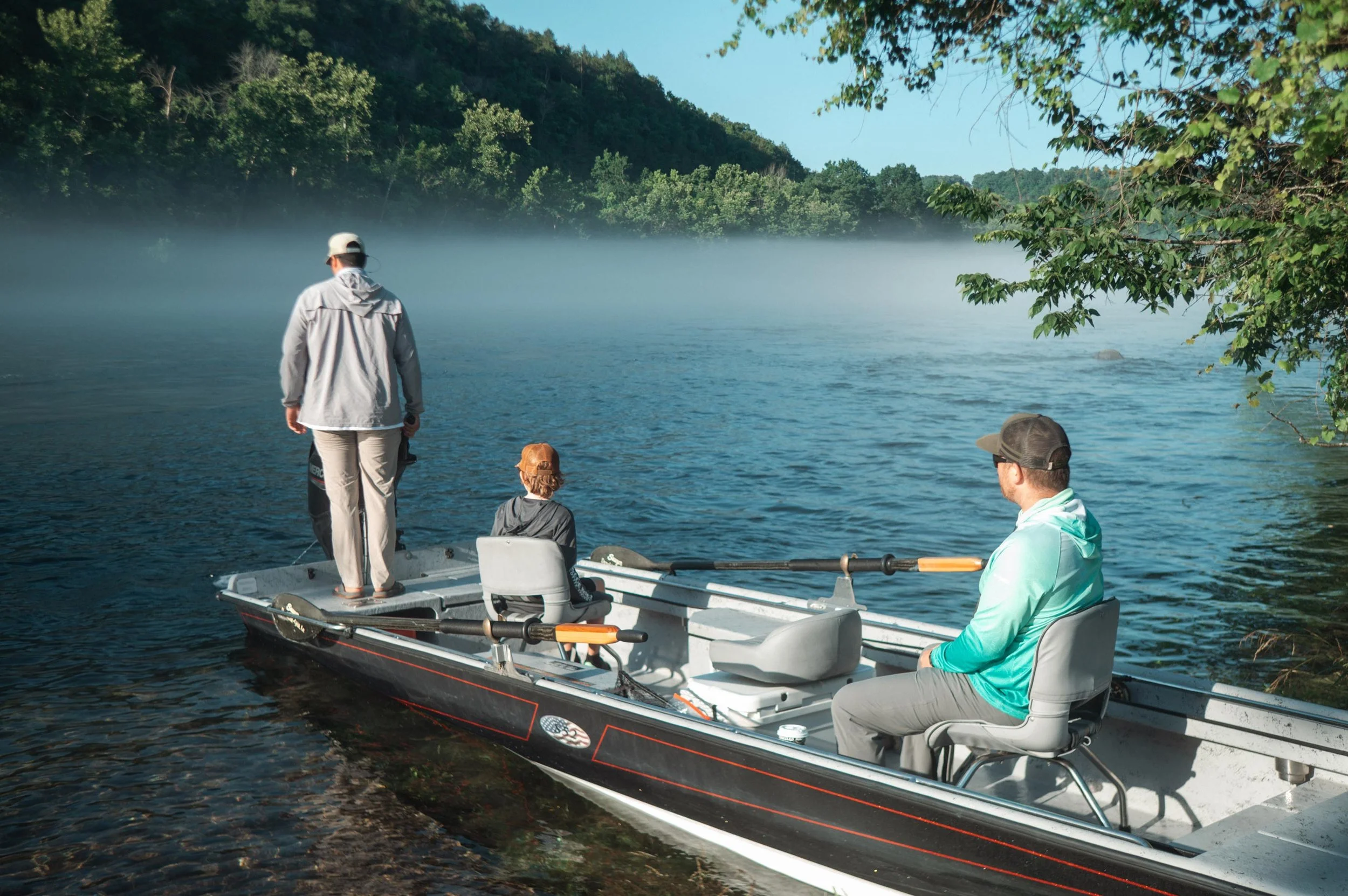 Four people are on a small fishing boat on a lake with mist rising from the water. The boat is near the shore with trees in the background. One person is standing at the back of the boat, holding a fishing rod, while the others sit or stand nearby.