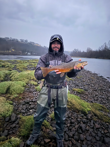 Man in rain gear standing on rocky riverbank holding a fish