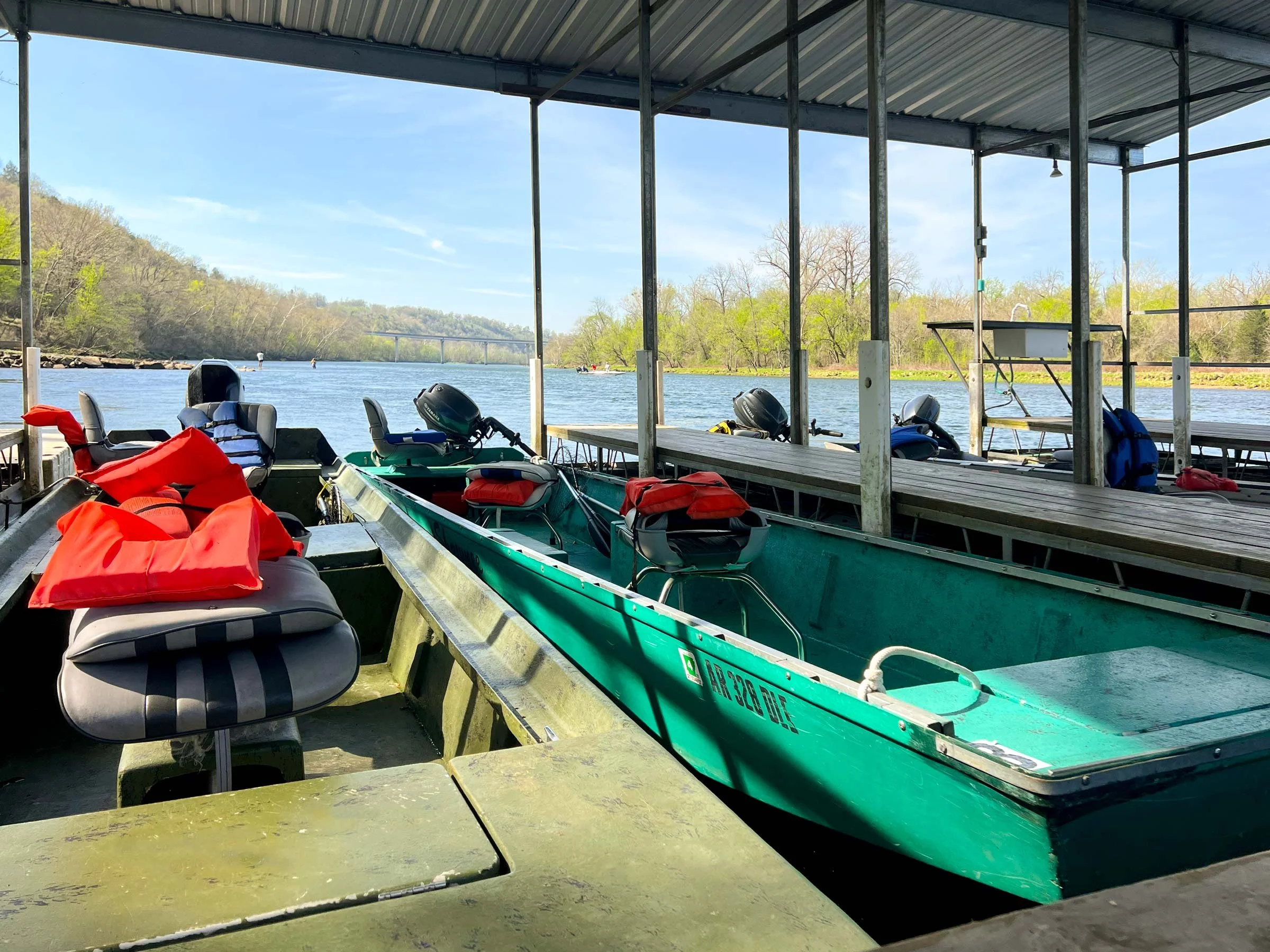 A scenic view of a river with a boat dock and kayaks, life jackets, and motors stored under a metal roofed dock. The river is calm, lined with trees and a distant bridge visible under a partly cloudy sky.