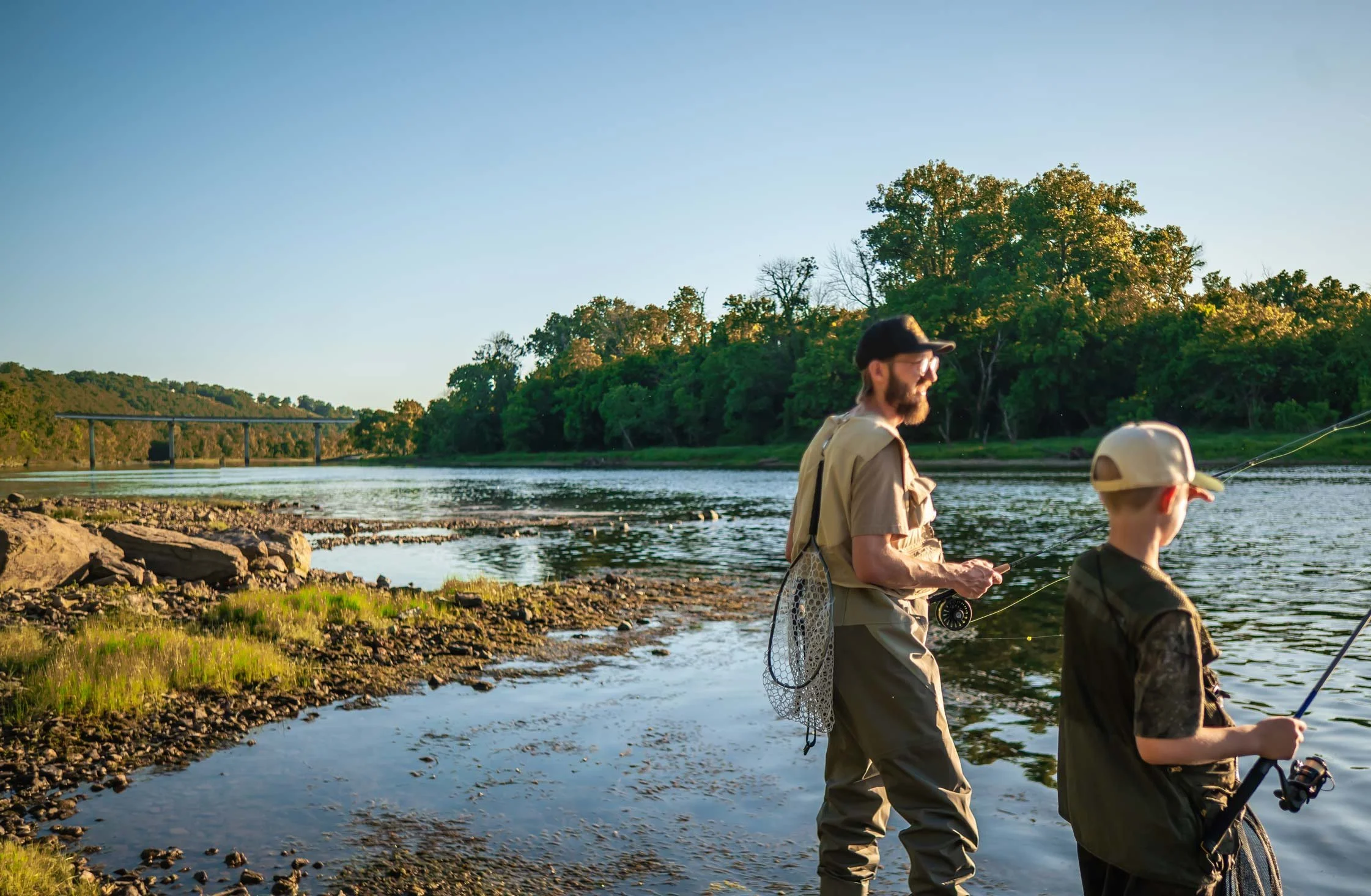 An adult and a child fishing by a river at sunset, with a bridge and wooded hills in the background.
