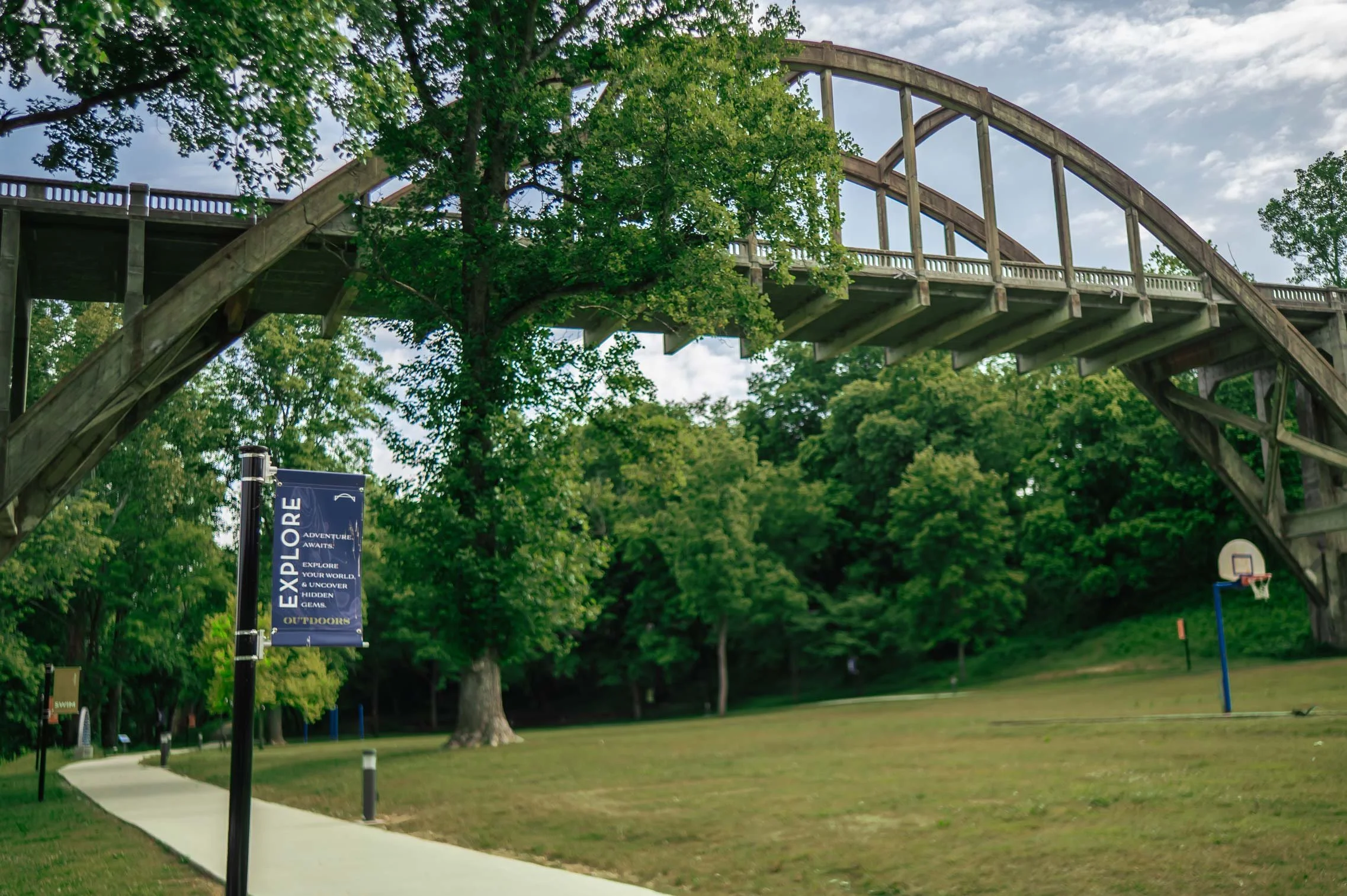 Wooden arched bridge over a park with green trees and cloudy sky, part of an outdoor recreation area with a basketball court and a sign that reads "Explore Outdoor".