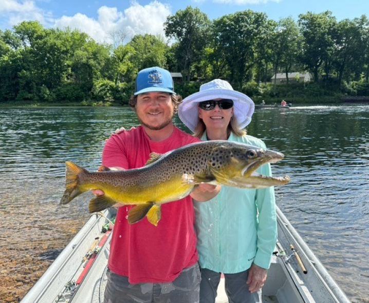 A young man and an older woman standing on a boat holding a large fish, with a river and trees in the background on a sunny day.