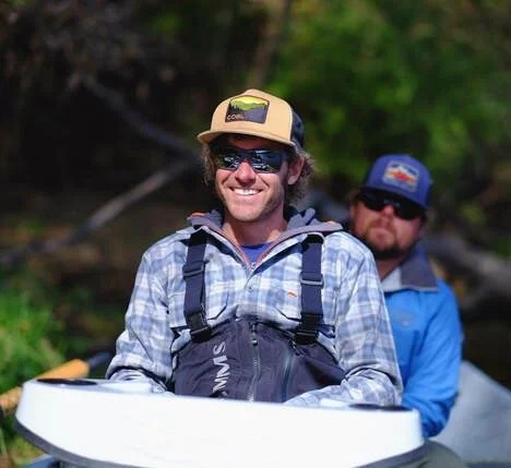 Two men in outdoor gear with life jackets, sitting in a canoe on a river surrounded by trees.