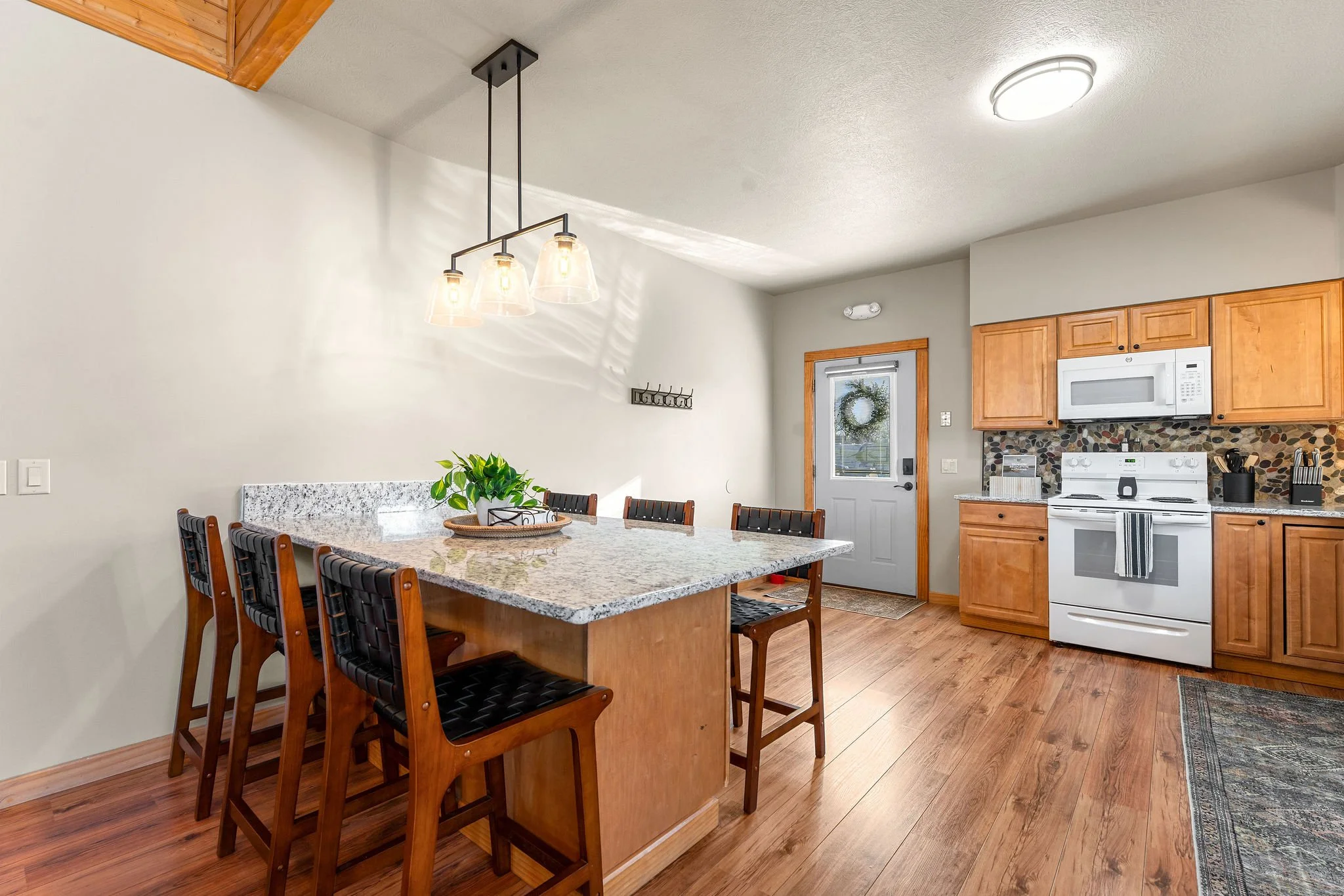 Kitchen with wooden cabinets, white appliances, and a granite island counter with seating. There's a door with a wreath, a hanging light fixture above the island, and hardwood flooring.