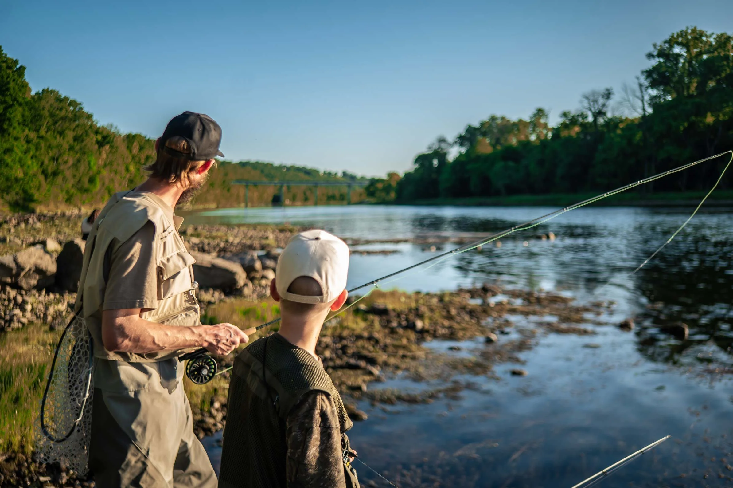 A man and a boy fishing by a river with trees in the background on a sunny day.