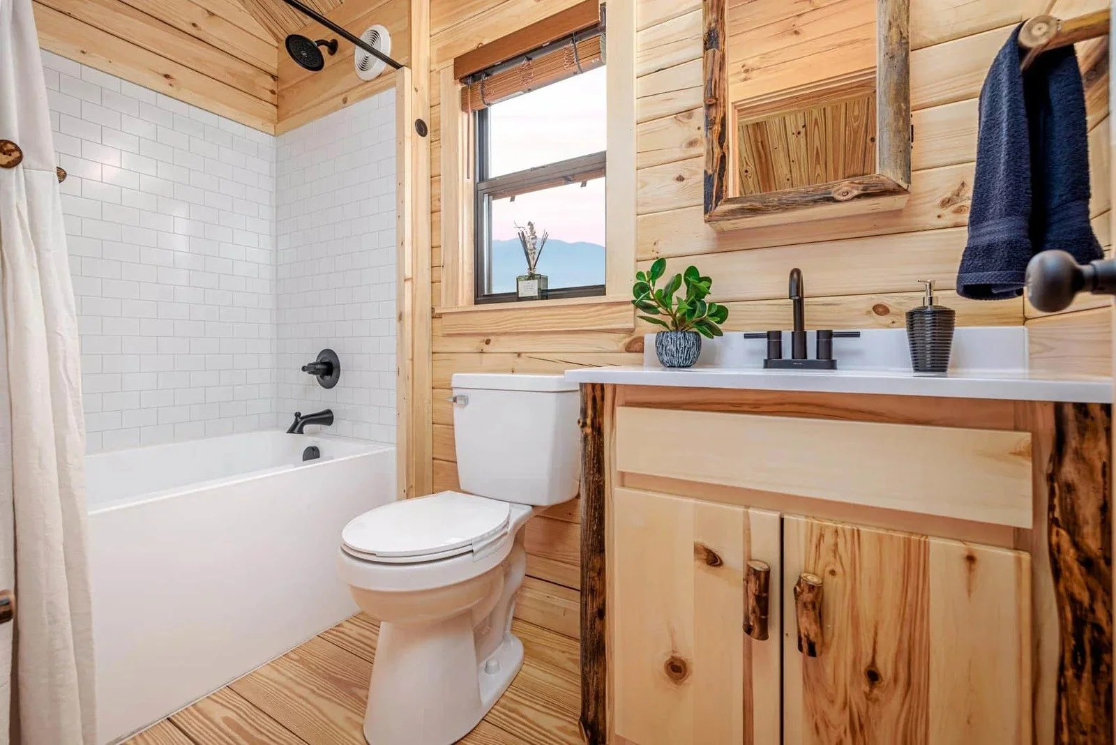 Bathroom with wood-paneled walls, a bathtub with black fixtures and white subway tile, a toilet, a wooden vanity with a white countertop, a black faucet, a plant, a black soap dispenser, a mirror, a window, and blue towels hanging on a towel rod.