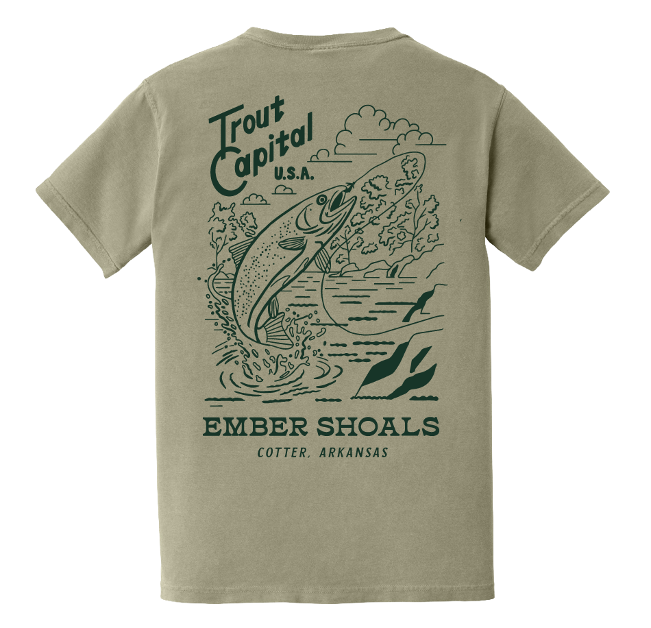 A beige T-shirt featuring a graphic of a trout leaping out of the water with trees and clouds in the background. Text reads 'Trout Capital U.S.A. Ember Shoals Cotter, Arkansas'.