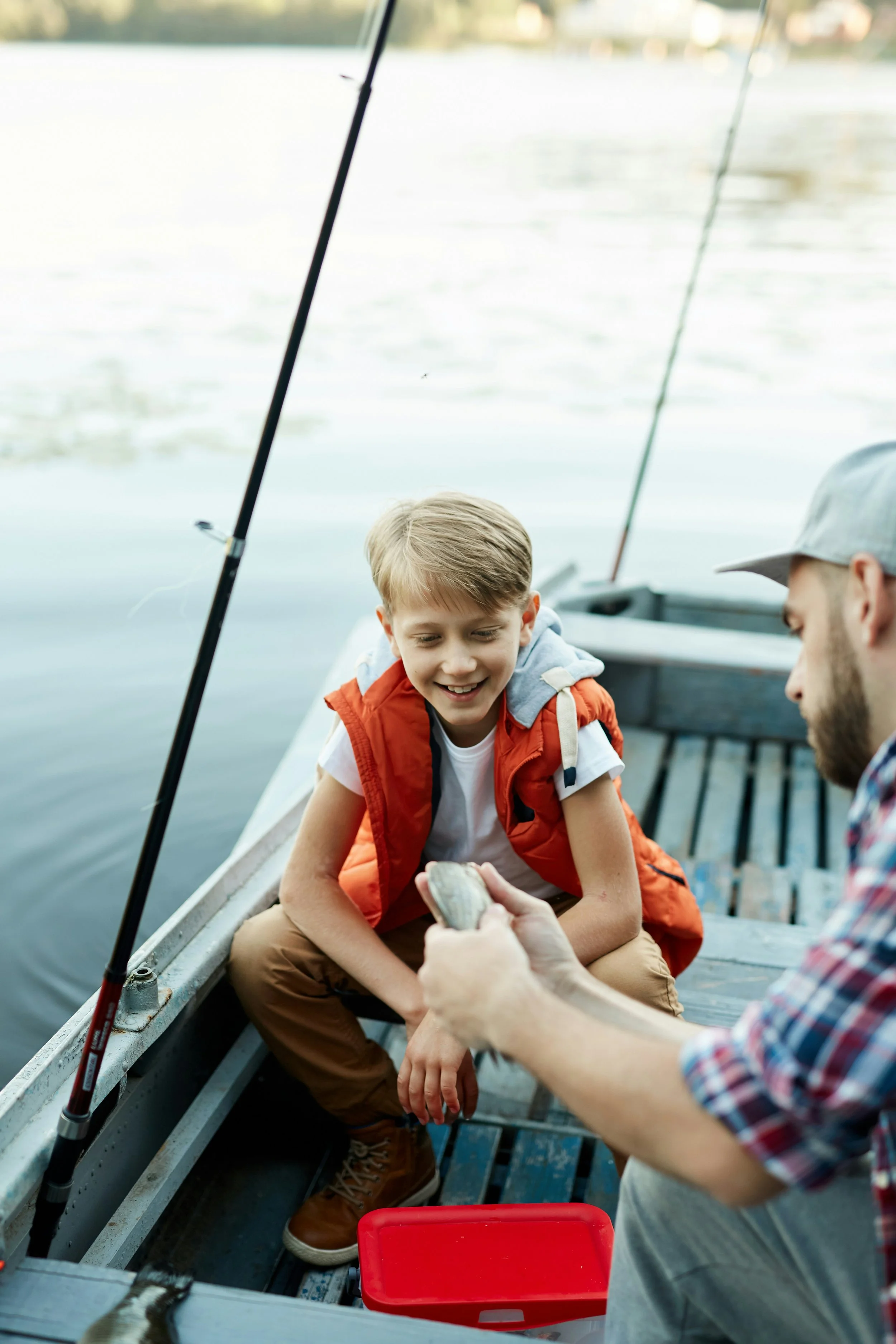 A young boy sitting in a boat, smiling and looking at an adult who is holding a fish, with fishing rods and water in the background.