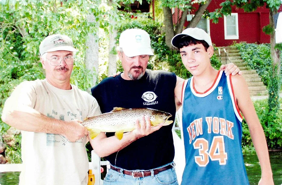 Three males outdoors, one holding a large fish, with trees and a house in the background.