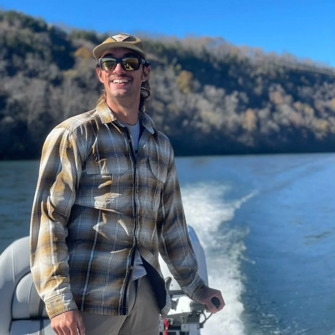 A man wearing sunglasses, a cap, and a plaid shirt is standing on a boat, smiling, with water and a forested hillside in the background.
