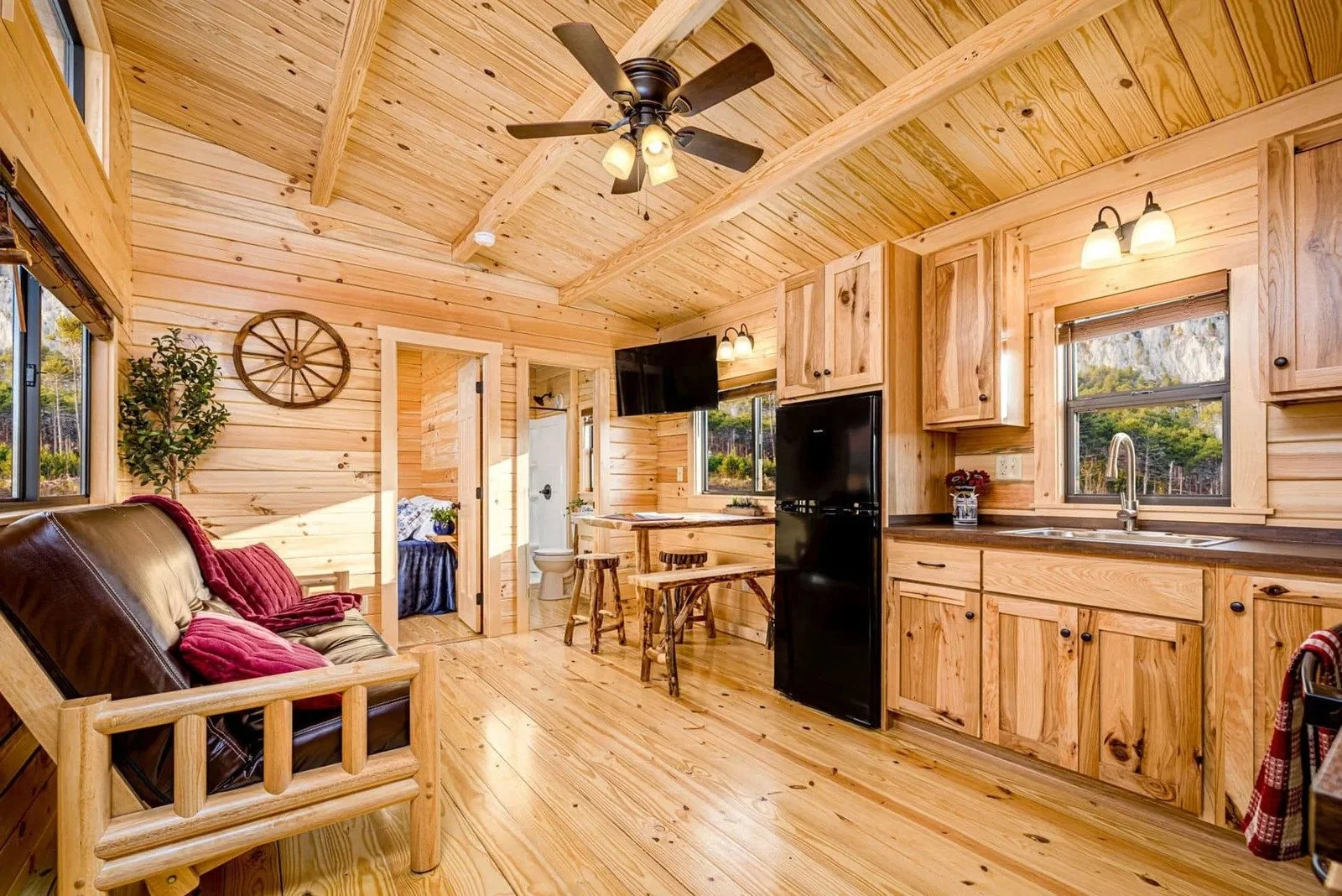 Cozy interior of a wooden cabin with a vaulted ceiling, sitting area with leather and wooden furniture, a small kitchen with wooden cabinets, a refrigerator, a sink, windows showing a scenic outdoor view, and a doorway leading to a bathroom.