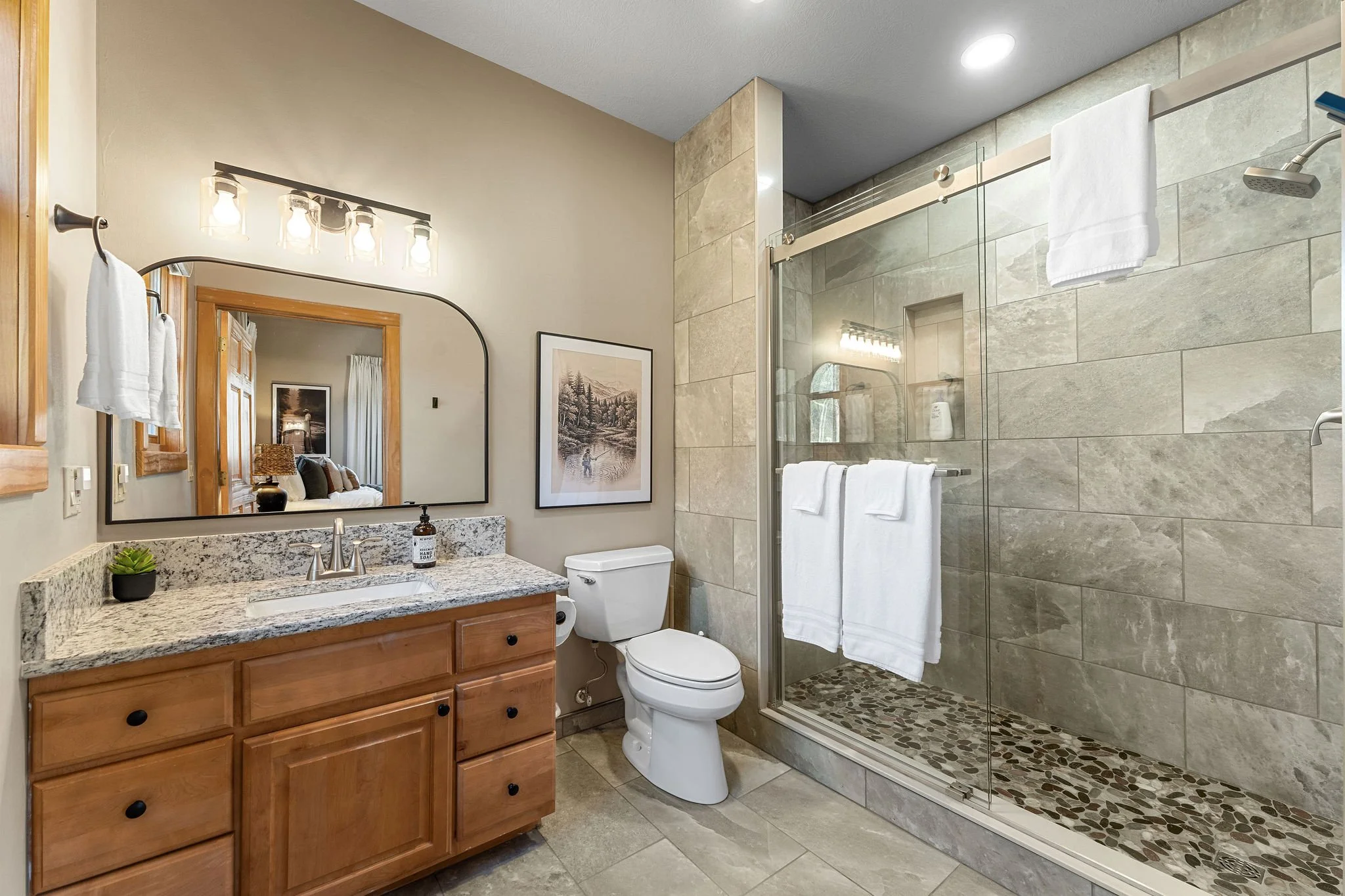 A modern bathroom with a wooden vanity and granite countertop, a mirror, a white toilet, and a glass-enclosed shower with beige tiles and pebble flooring, with towels hanging on the wall.
