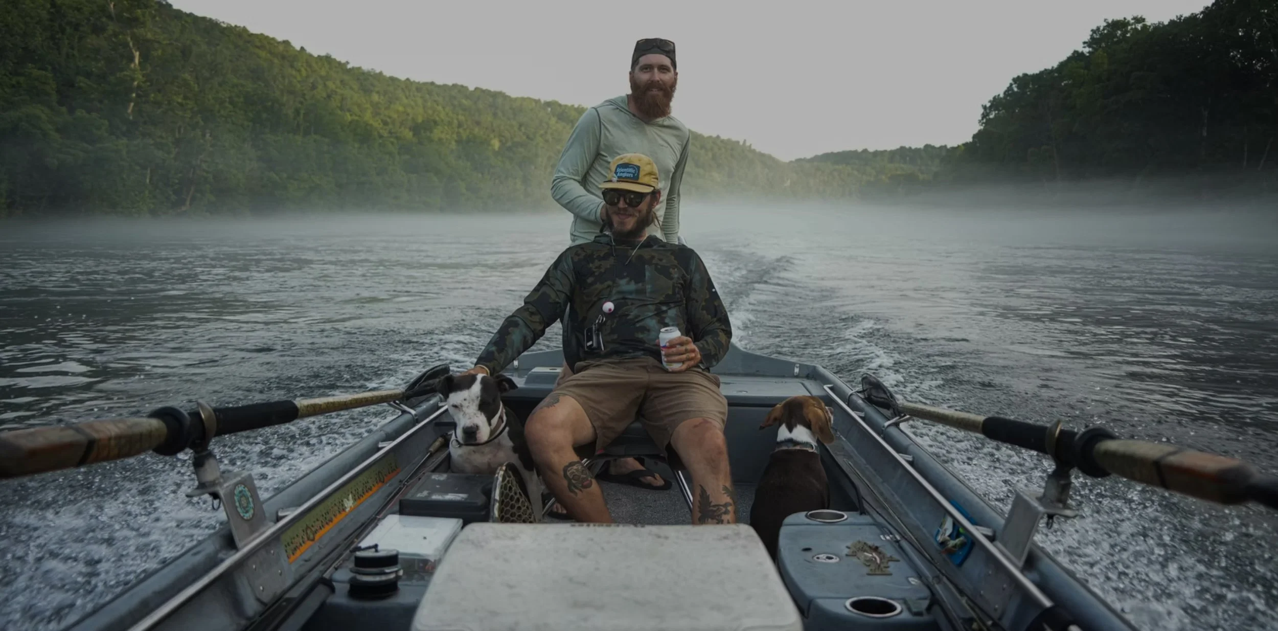 Two men and two dogs on a boat on a river with green forests on both sides, one man with a beard standing and another sitting holding a drink, both wearing casual outdoor clothing and sunglasses, with a misty river and trees in the background.