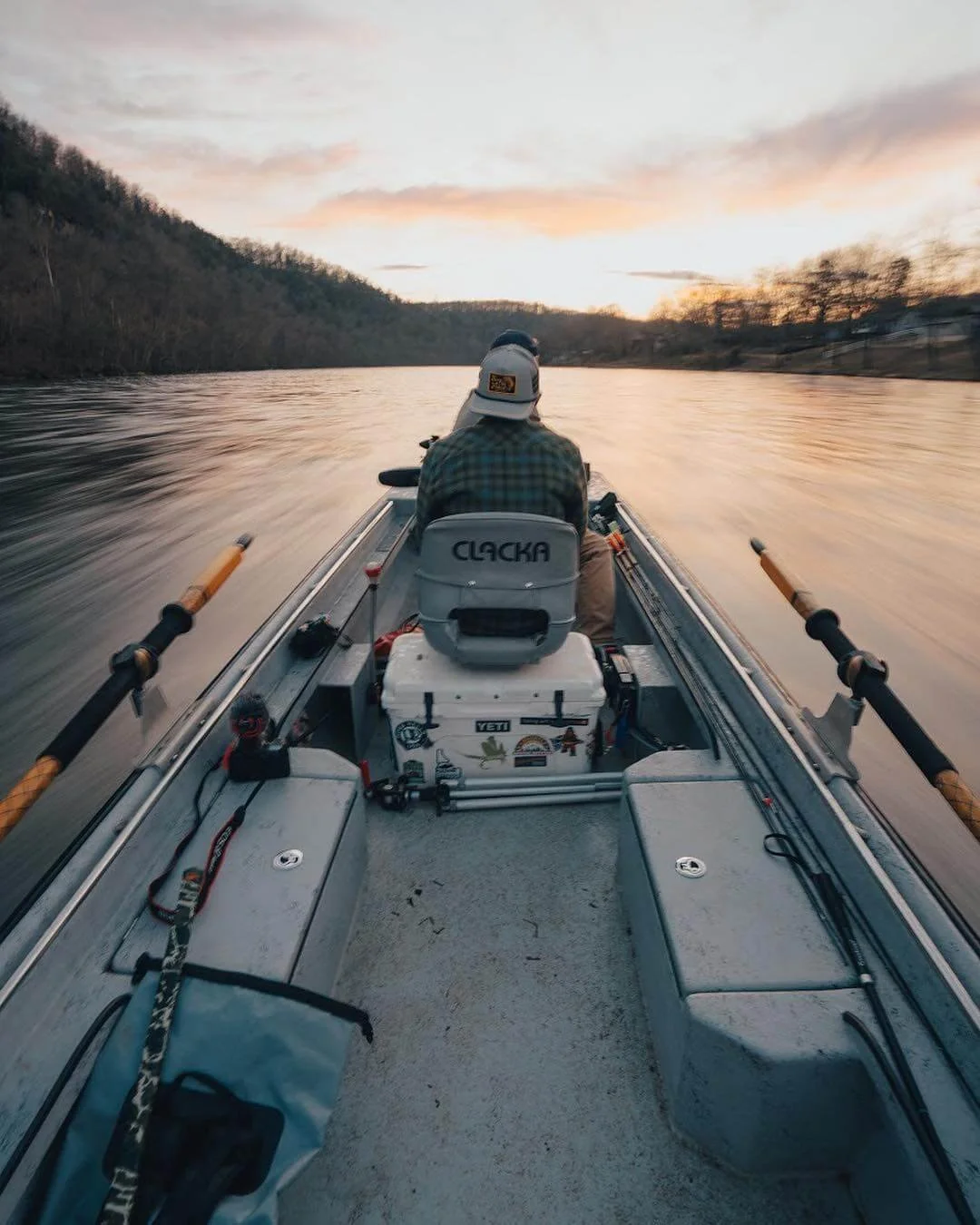 Person fishing from a boat on a river at sunset, with trees on the riverbanks and fishing gear visible.