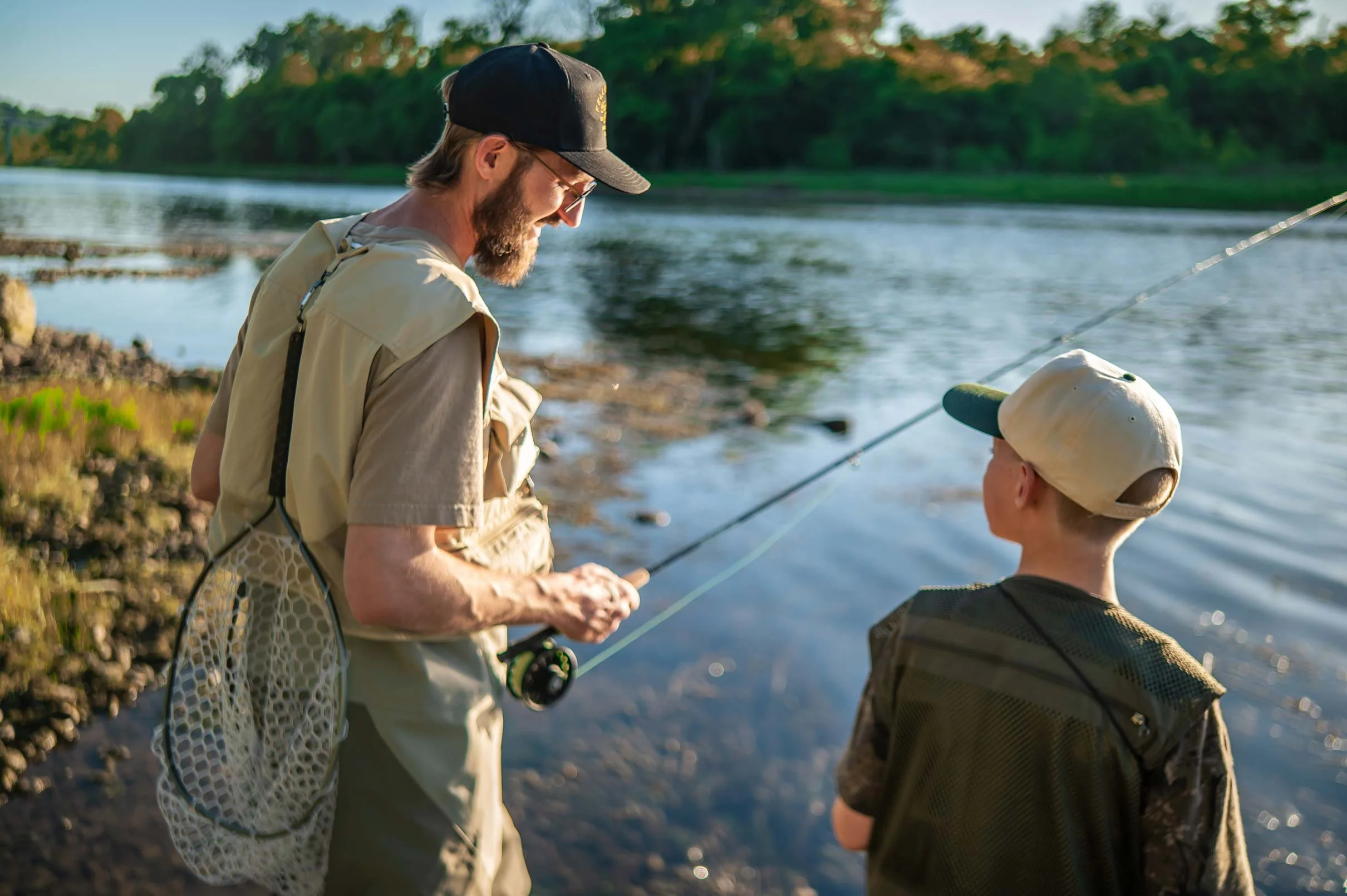 An adult man and a young boy fishing together by a river during late afternoon or early evening. The man is wearing a beige fishing vest and a black cap, while the boy is wearing a dark shirt and a light-colored hat. They are both smiling and looking