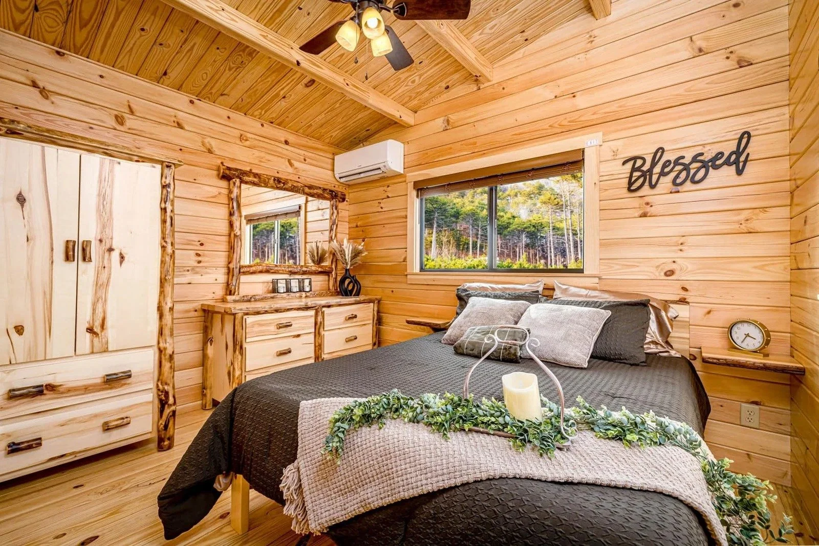 A cozy bedroom with wooden walls, a dark bedspread, decorative pillows, a mirror, a dresser, a window showing a forest view, and a sign that says 'Blessed' on the wall.