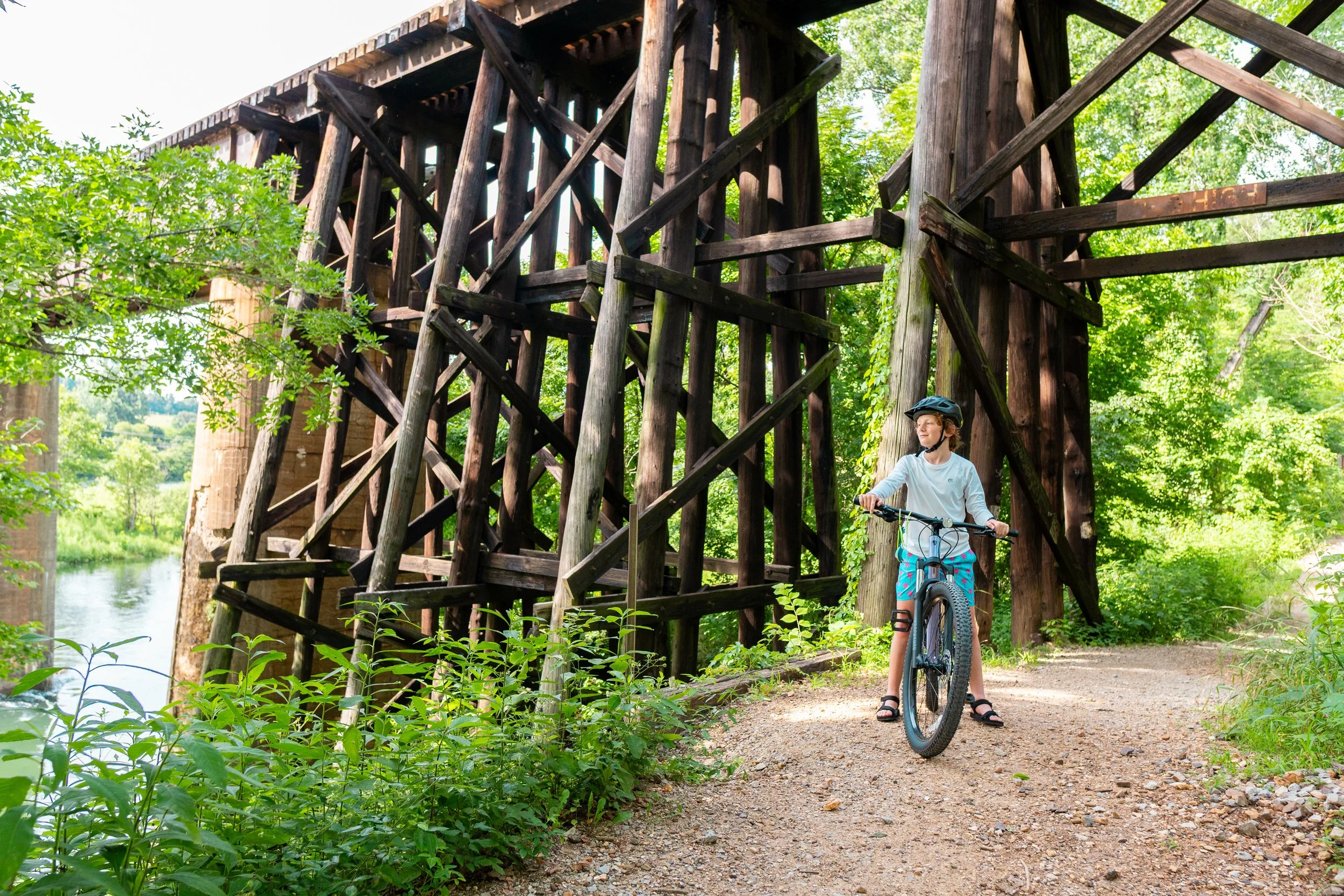 A young girl wearing a helmet and riding a bicycle under a large wooden trestle bridge surrounded by green trees and foliage.