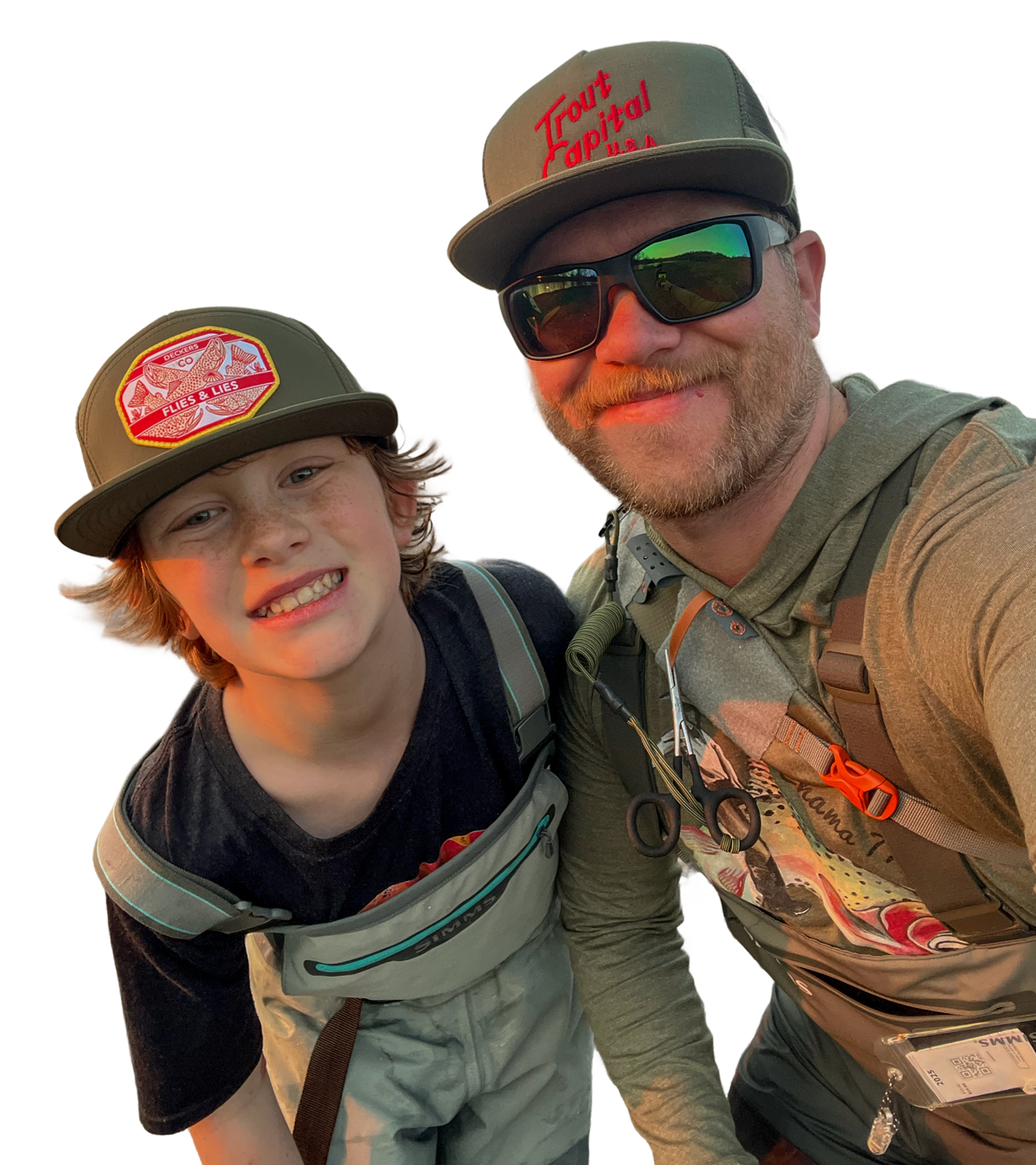 A man and young boy outdoors, smiling and taking a selfie, wearing hats and casual outdoor clothing, with backpacks and fishing gear.