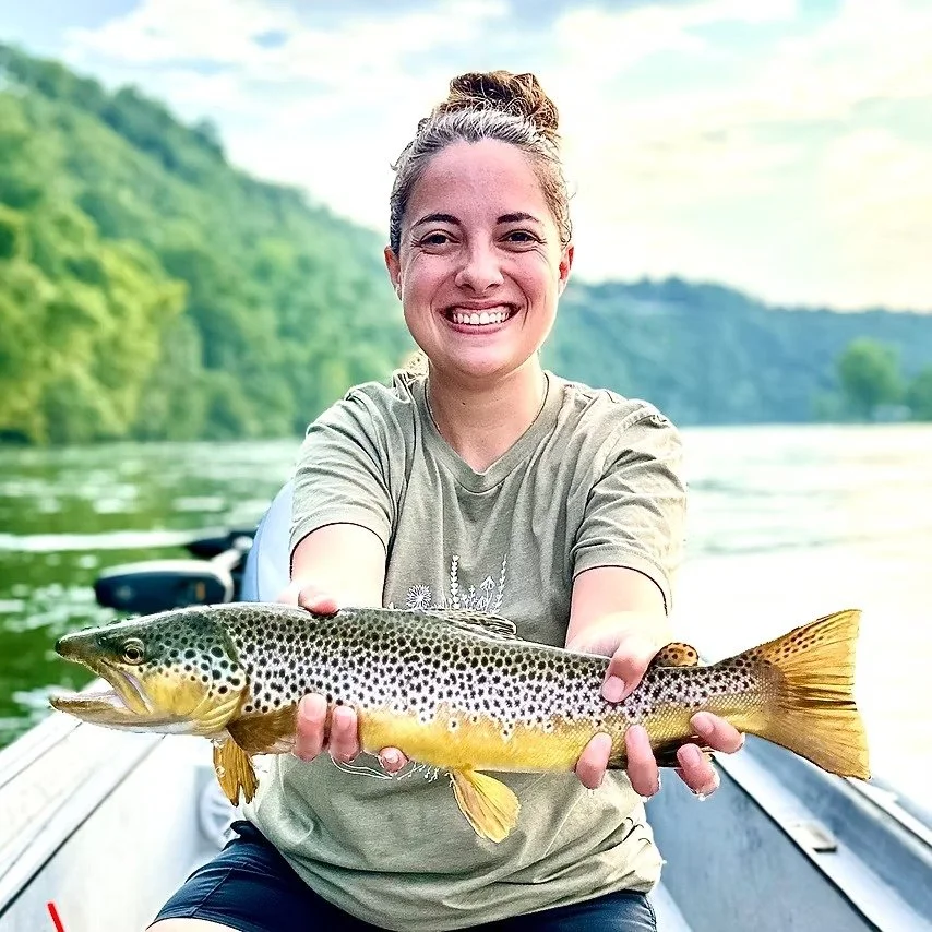 Woman holding a large fish on a boat in a river with green hills in the background.
