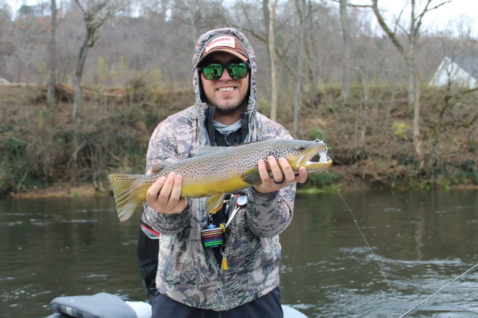 Man in camouflage jacket and cap holding a large rainbow trout by a riverbank, smiling at the camera with trees and a house in the background.