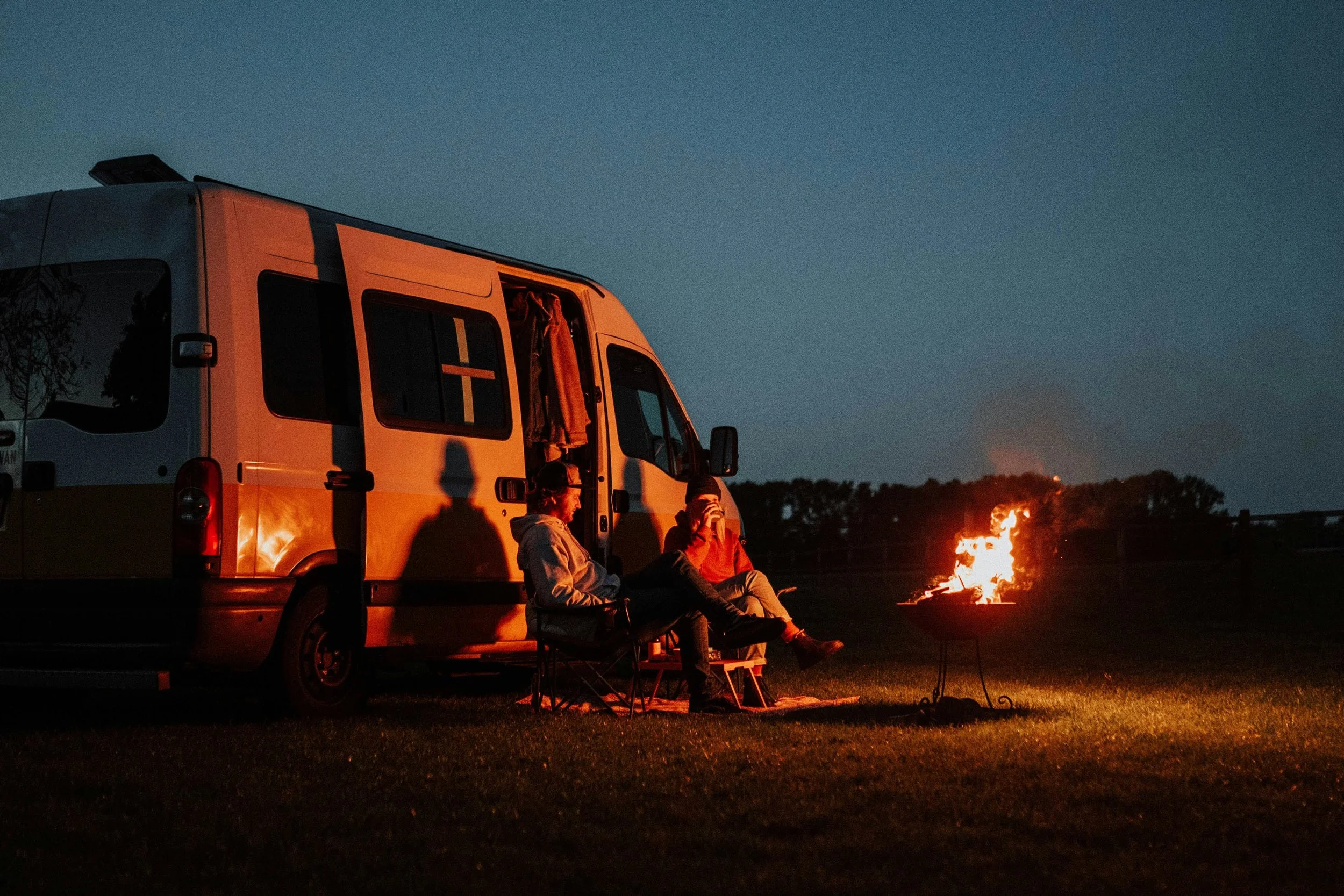 People sitting near an orange and white van with a campfire in the foreground at dusk or night.