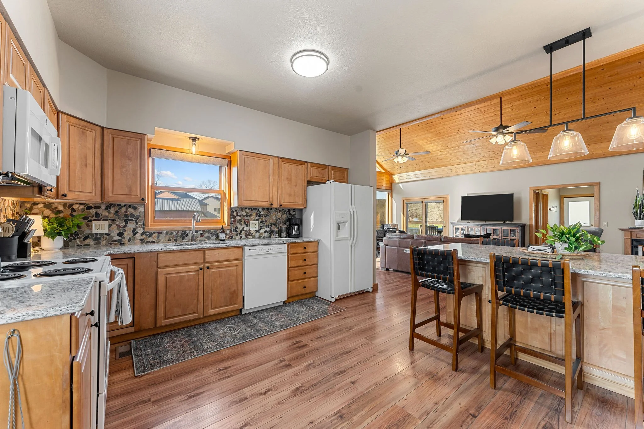 An open-concept kitchen and living room area with wooden cabinetry, a white refrigerator, a stove, a granite countertop, a window, ceiling fans, a television, a fireplace, and a wooden dining table with chairs.