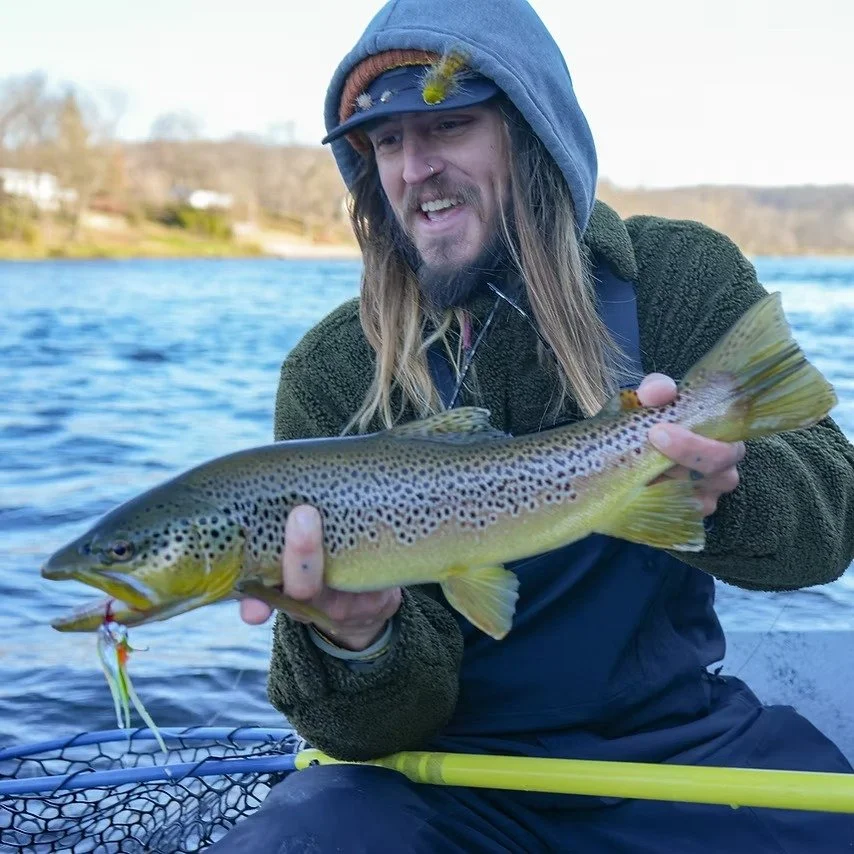 A man with long hair, a hoodie, and a cap, smiling and holding a large rainbow trout over water.