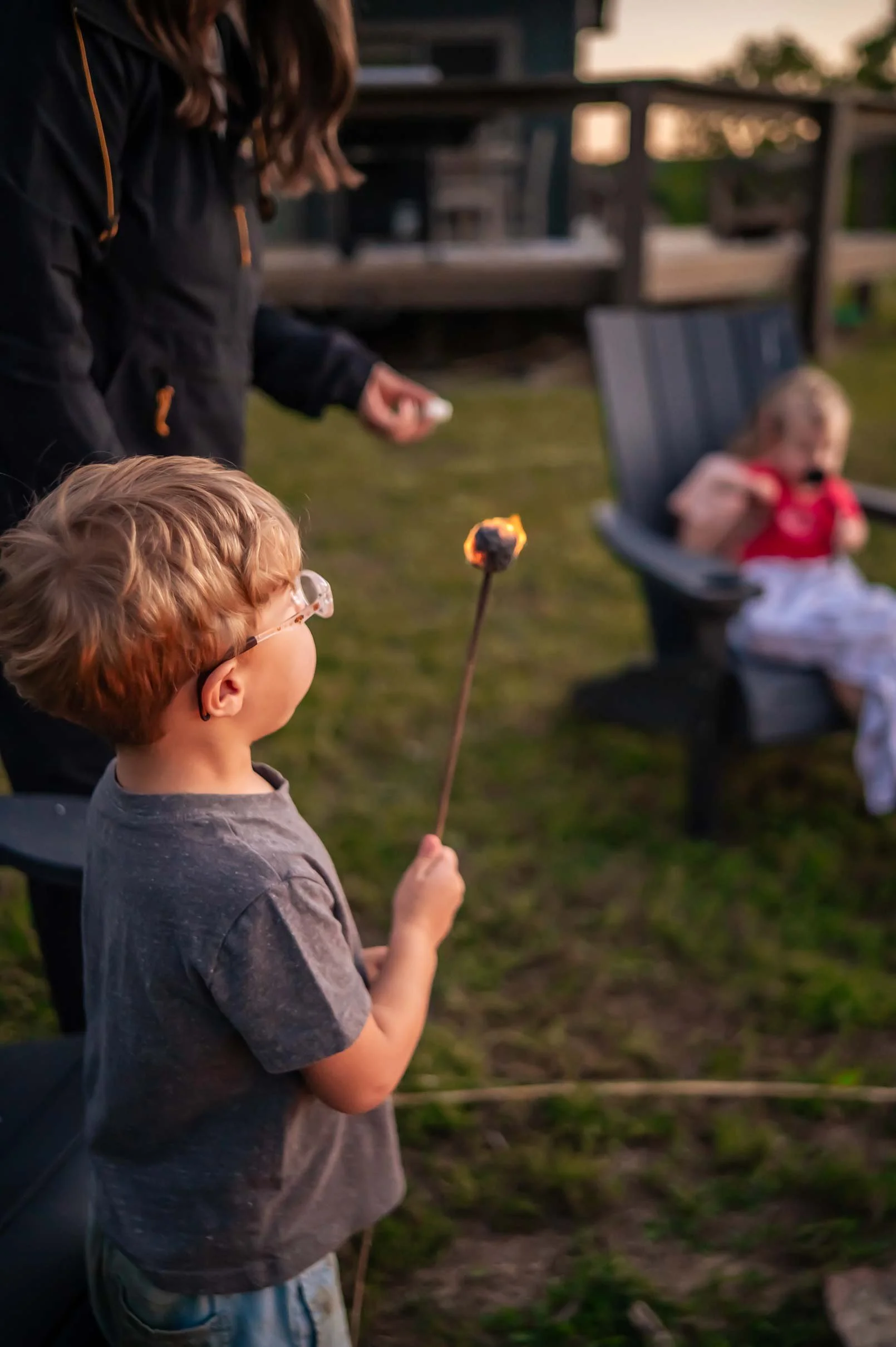 A boy with glasses holding a lit stick of marshmallow over a fire, with a woman and a girl sitting on a bench in the background during sunset.