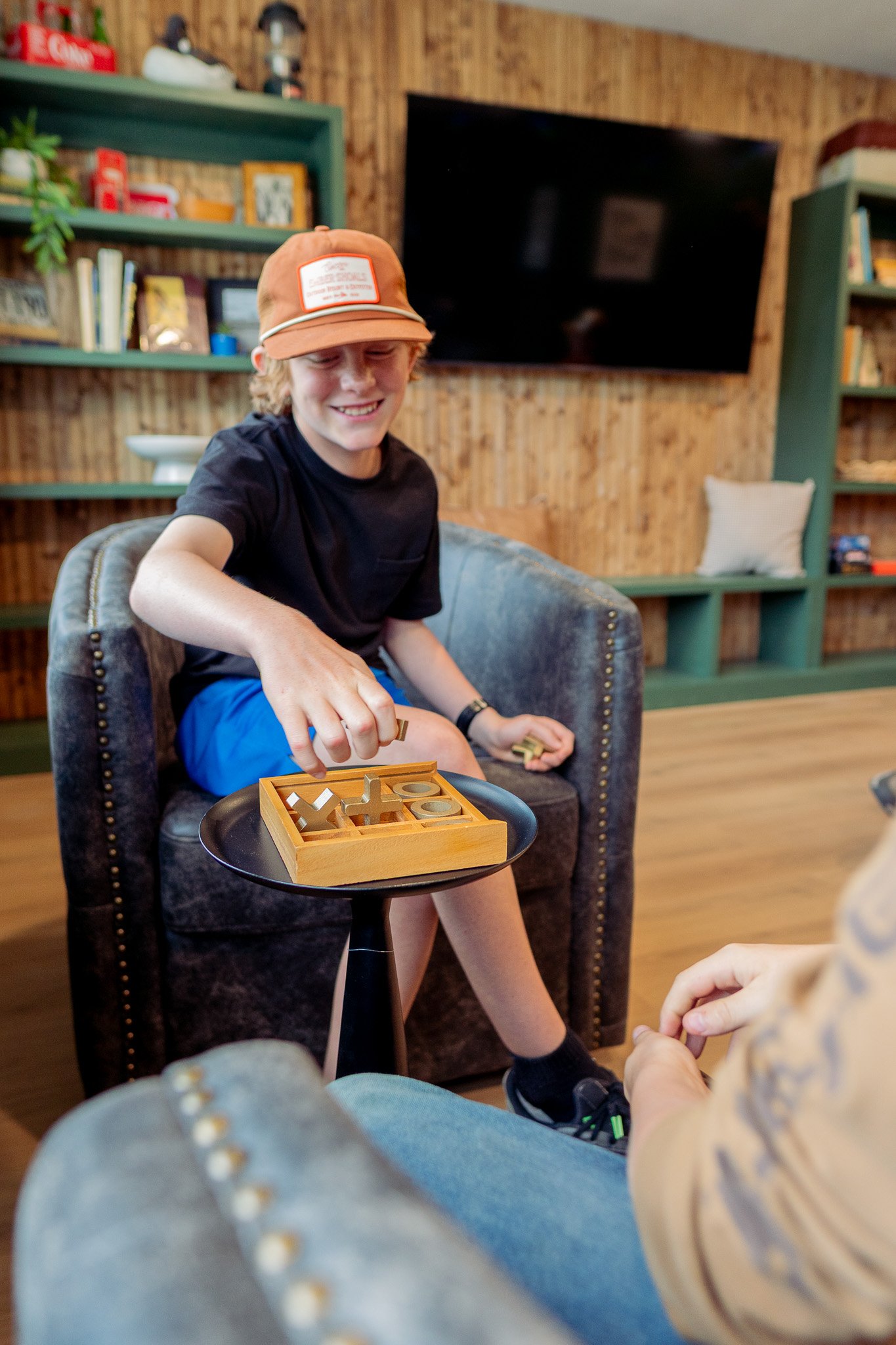 A young boy smiling and playing a board game with a rack of letter tiles, sitting on a charcoal armchair in a cozy living room with wood-paneled walls and green bookshelves.