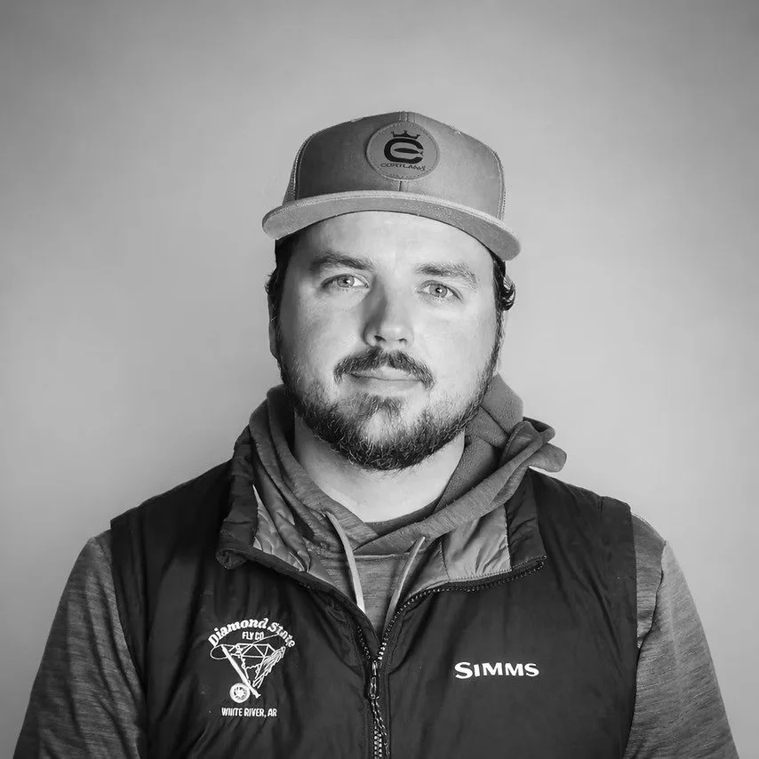 A man with a beard and mustache wearing a baseball cap, hoodie, and a vest with logos, looking at the camera against a plain background.