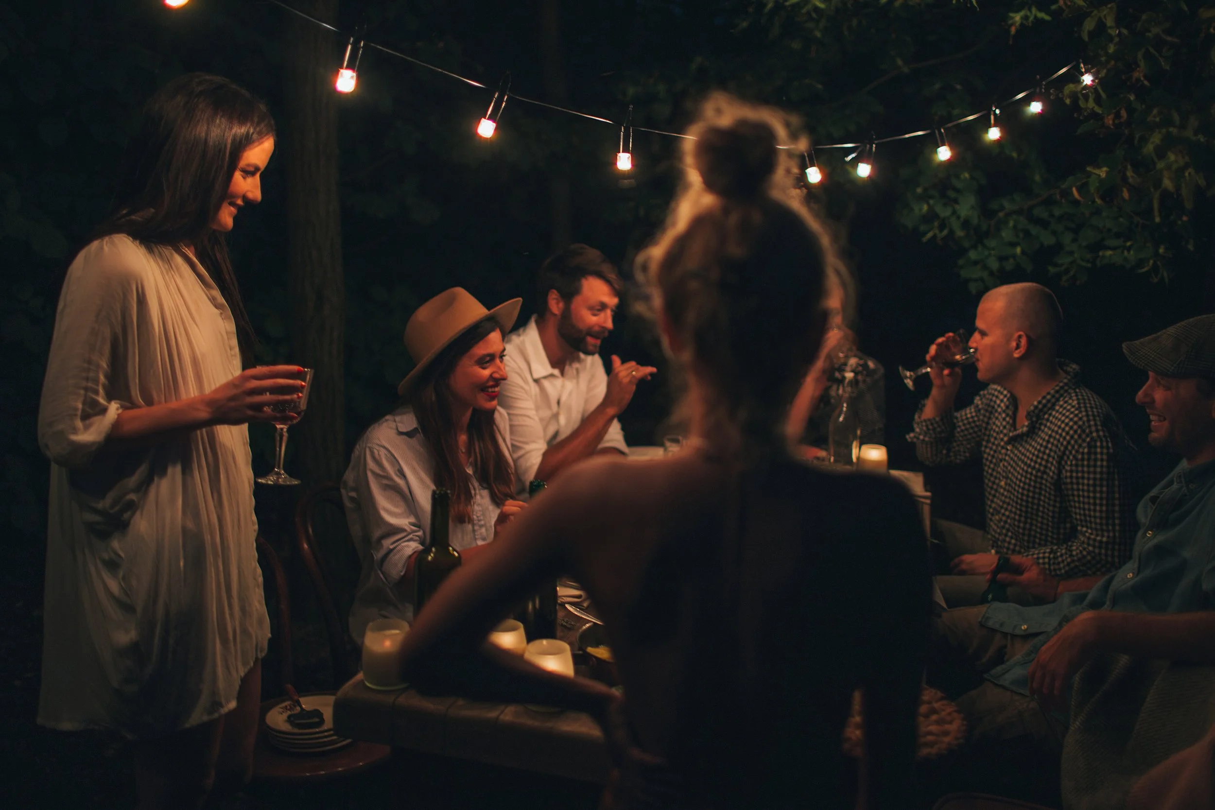 People enjoying an outdoor evening gathering with string lights overhead, some sitting at a table with drinks and food, and a woman standing with a glass of wine.