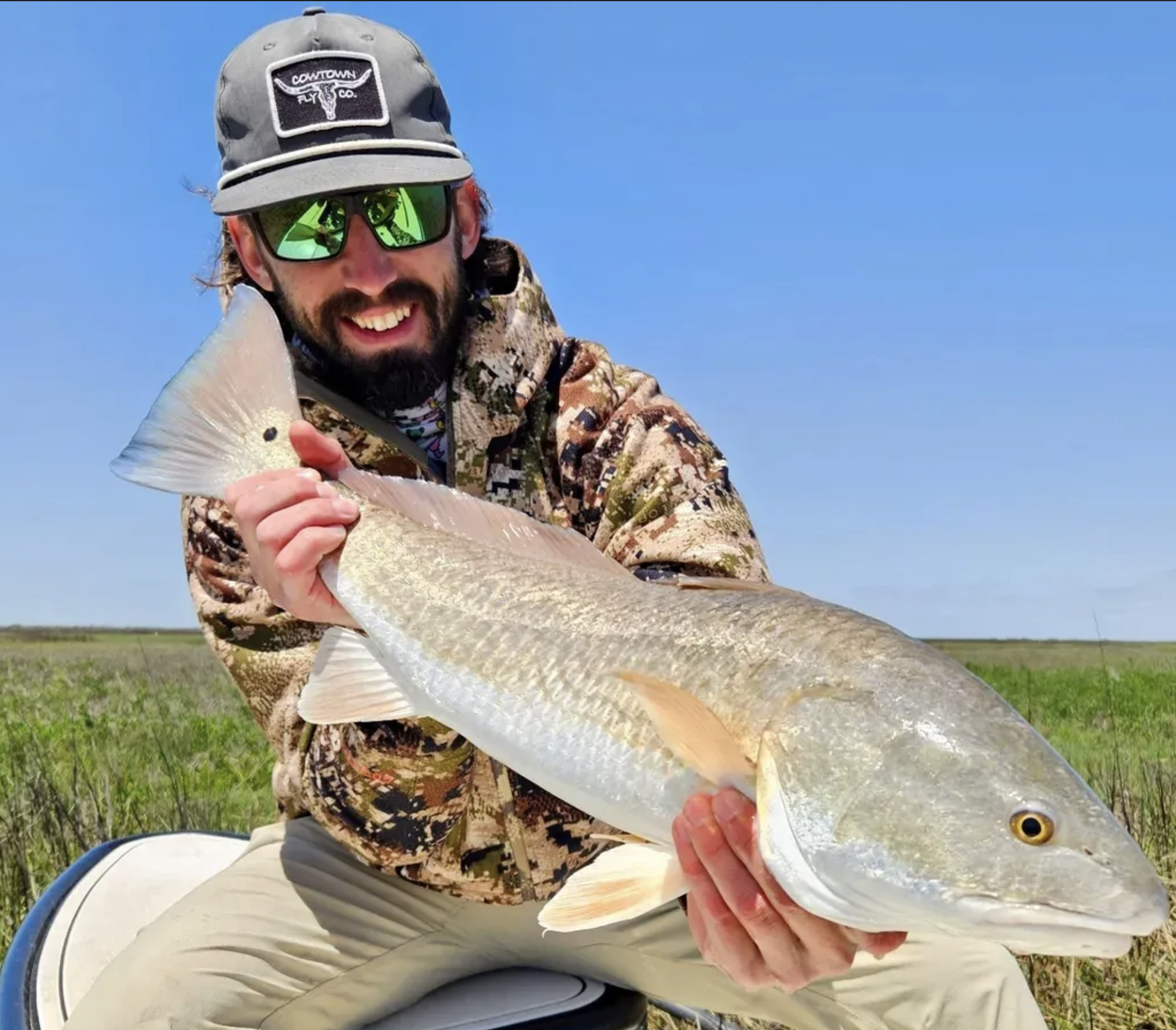 Man in camouflage jacket and gray cap holding a large fish on a boat in a grassy wetland area under a clear blue sky.