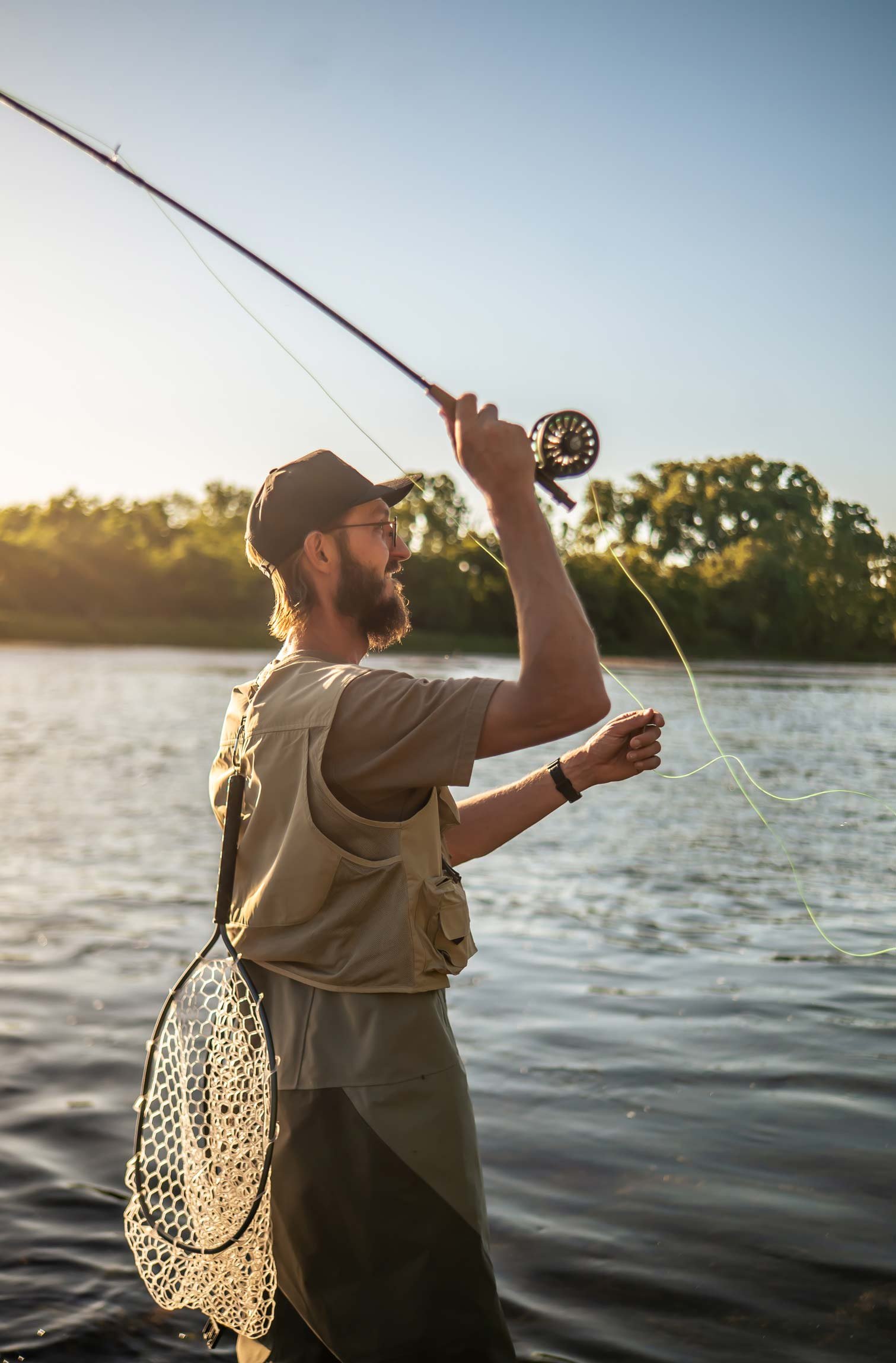 A man with glasses and a beard fishing with a fly rod in a water body during sunset, wearing a cap, beige shirt, and fishing vest, with a fishing net attached to his waist and trees in the background.