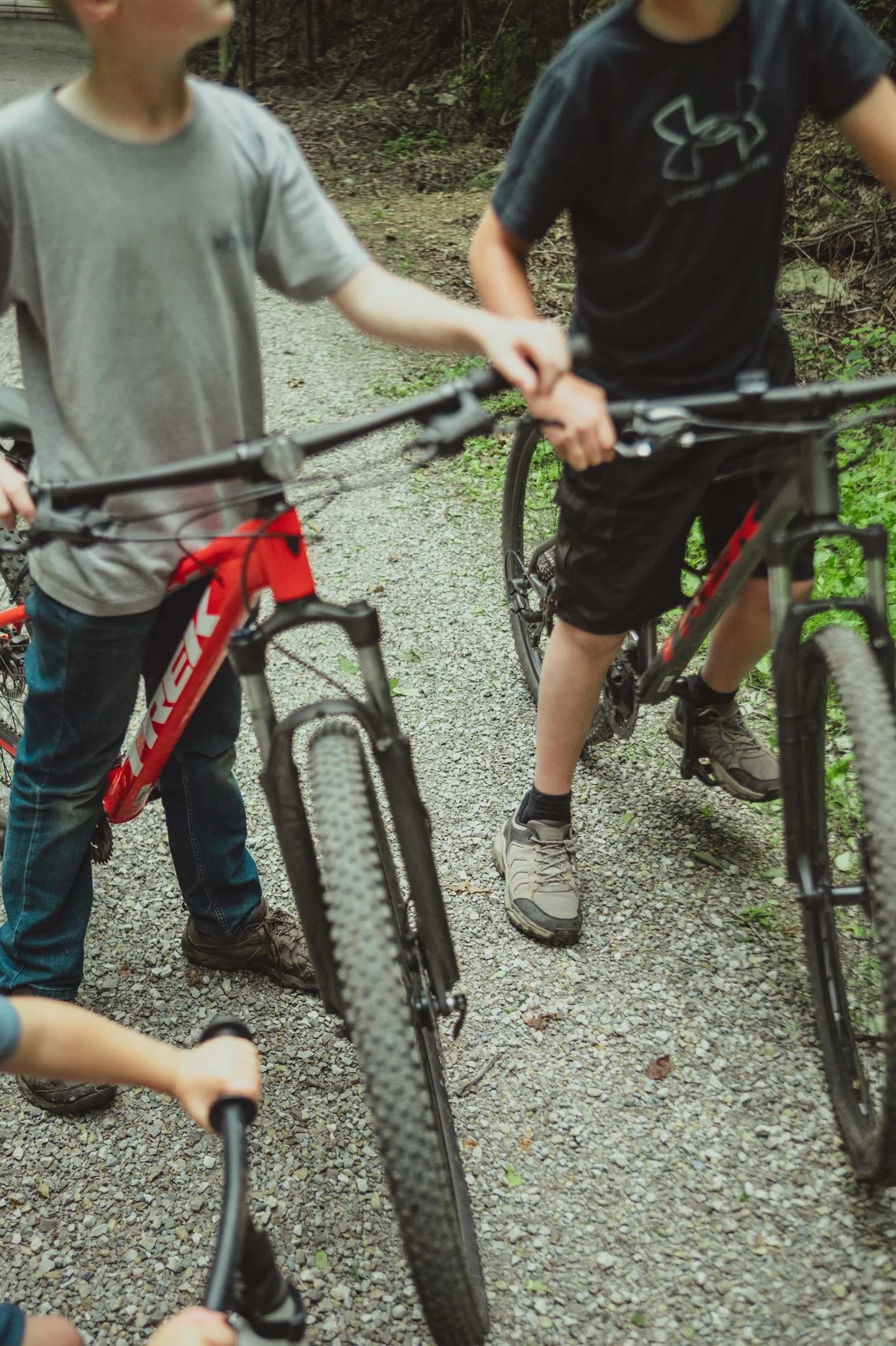 Two children are holding onto their mountain bikes on a gravel trail in a wooded area.