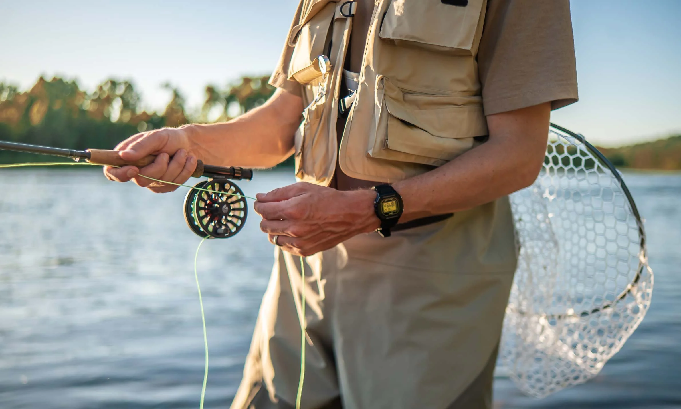 A person fishing in a body of water during daylight, wearing a beige fishing vest and holding a fishing rod with a reel. A fishing net is hanging from their waist.