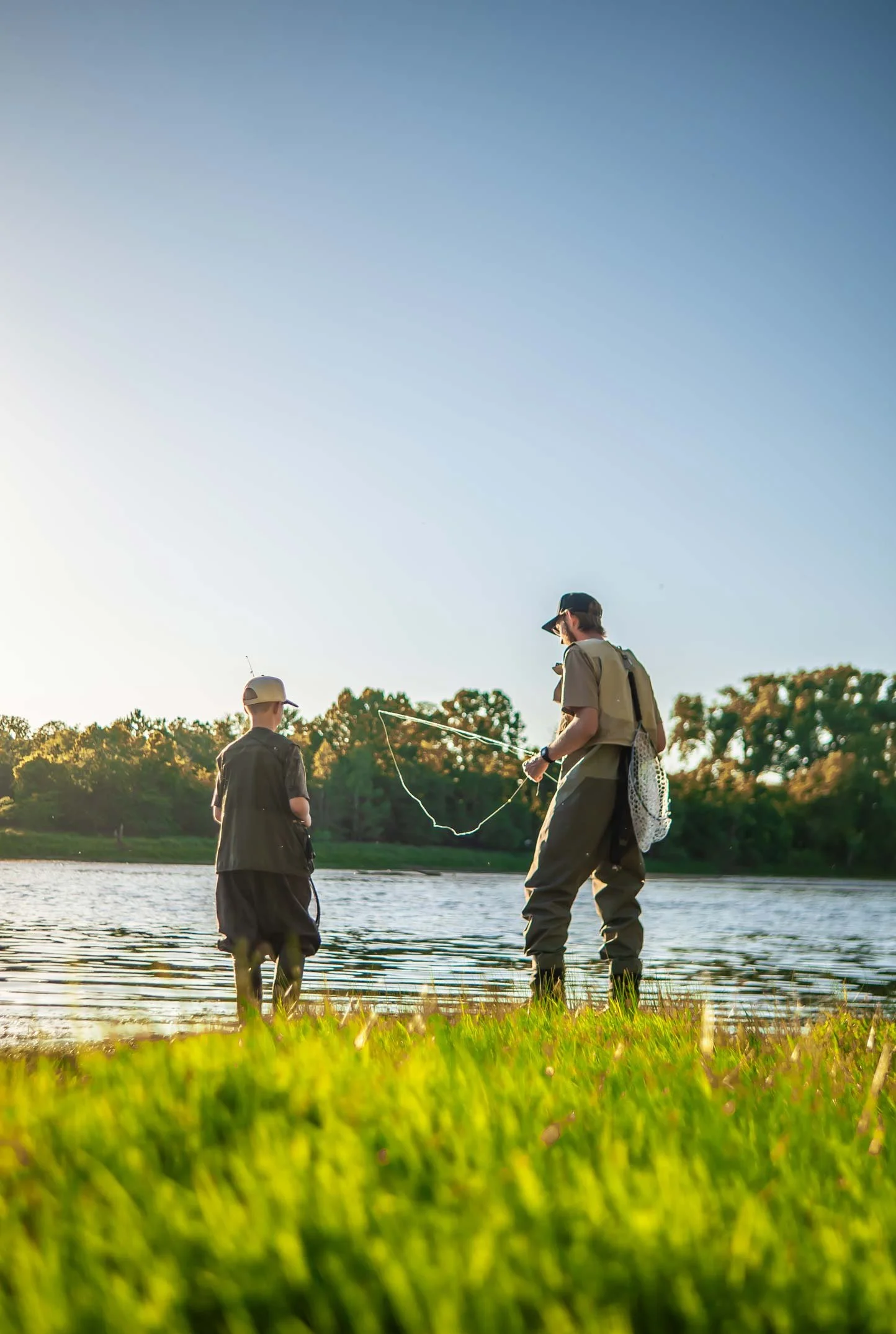Two people, likely a child and an adult, fishing by a lake during sunset or early evening, with trees in the background and green grass in the foreground.