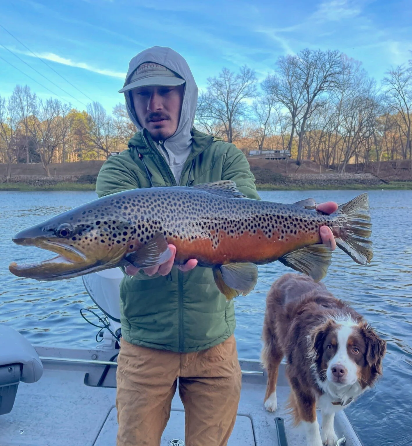 A man in outdoor clothing holding a large fish, an Australian shepherd dog with brown and white fur looking at the camera, on a boat by a lake with trees in the background.