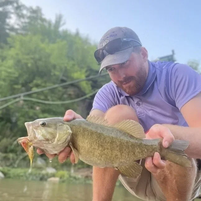 A man wearing a baseball cap, sunglasses on his head, and a purple shirt, holding a large fish with a smaller fish in its mouth, near a body of water with green trees in the background.