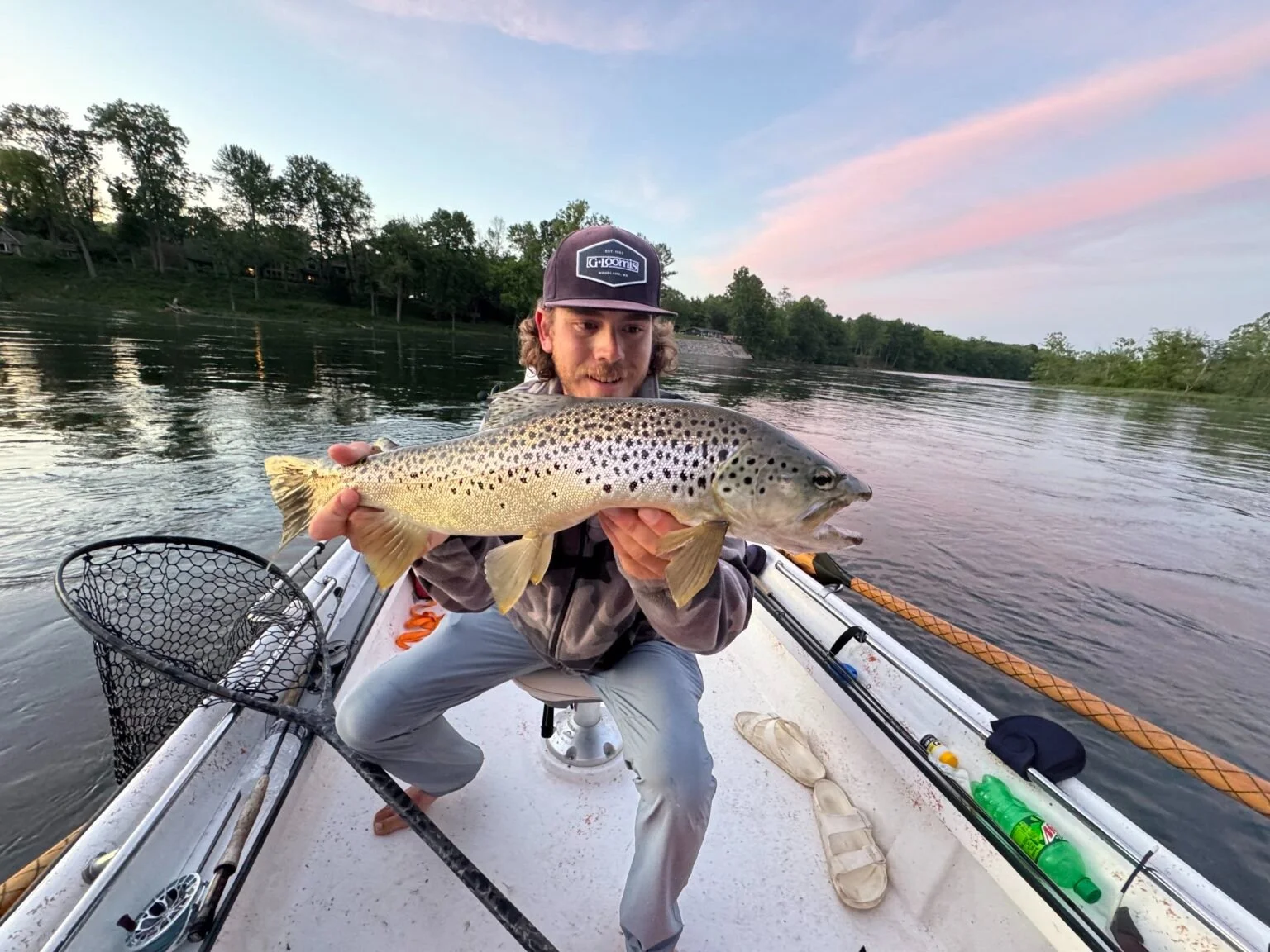 A man in a boat holding a large fish, likely a trout, with a scenic river and sunset in the background.